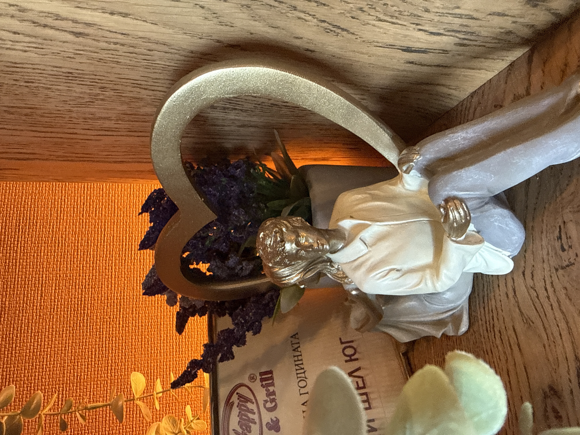 A decorative figurine, possibly an angel or a similar spiritual figure, is prominently displayed in this indoor scene, rotated ninety degrees clockwise from its natural orientation. The figure has a golden head and hands, and wears a white upper garment and a grey lower robe, kneeling or sitting. Behind it stands a large, golden, heart-shaped arch or frame. Artificial purple flowers, resembling lavender, along with green foliage, are arranged around the figurine and within the heart shape, adding a natural, decorative touch.
The entire arrangement rests on a wooden surface, likely a shelf or table. To the left, a textured orange wall provides a warm background, possibly illuminated by artificial light, creating a soft glow. Another wooden surface is visible above and to the right, suggesting a wooden paneling or another shelf. In the bottom right foreground, a light-colored, artificial plant with small leaves is partially visible.
Beneath the figurine, a menu or brochure is visible, containing text in Cyrillic script. The legible text includes "Happy & Grill", indicating a restaurant or cafe. Further text reads "ГОДИНАТА" (GORDINATA), meaning "the year" in Bulgarian, and "ШЕЛ ЮГ" (SHEL YUG), translating to "Shell South". These details, combined with the location context of Radinovo, Bulgaria, confirm an interior setting within a Bulgarian establishment. The scene conveys a peaceful and decorative ambiance, typical of a dining or hospitality environment.