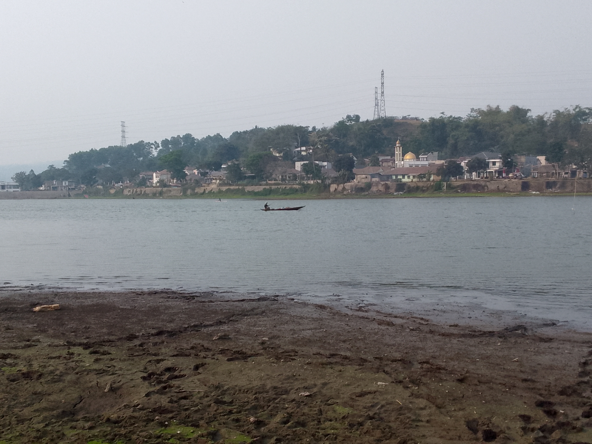 The image shows a body of water with a muddy shoreline in the foreground. In the middle ground, a small boat with a single occupant is being rowed across the water. In the background, there is a hillside with trees and a village of houses, including a building with a gold dome, suggesting it may be a mosque. Two large electrical towers are visible on the top of the hill, and power lines stretch across the sky. The overall atmosphere is hazy and muted, indicating an overcast or smoggy day. The scene is likely Ciburuy, Indonesia, given the presence of a mosque and the tropical vegetation.