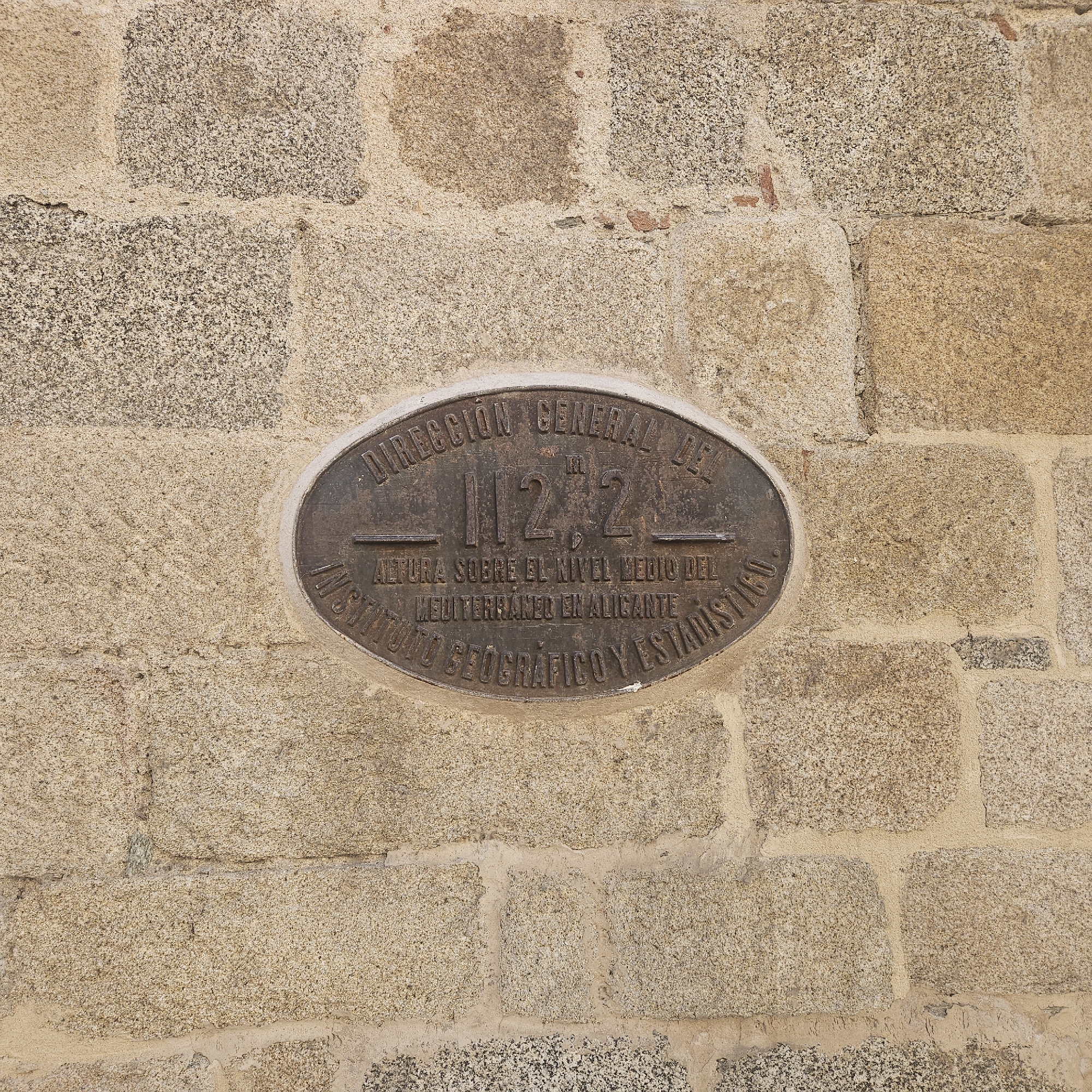 The image shows an oval, bronze-colored plaque affixed to a rough stone wall, likely part of a building. The plaque bears the inscription "Dirección General del 112,2 m. Altura Sobre el Nivel Medio del Mediterráneo en Alicante, Instituto Geográfico y Estadístico". The inscription is in Spanish. The wall is constructed of irregular, light-colored stone blocks joined with mortar. The overall scene suggests an official marker indicating the altitude above sea level, placed by the Spanish Geographical and Statistical Institute in reference to the average Mediterranean sea level in Alicante. The location is in the city of La Jonquera, Spain.