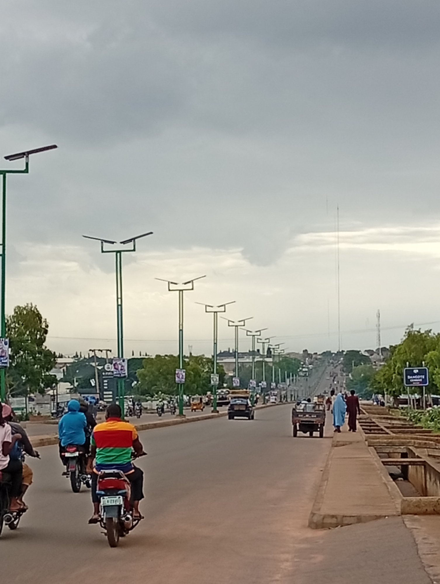 The image depicts a street scene in the city of Wuro Dole, Nigeria, on an overcast day. The road is a two-lane asphalt surface with a concrete pedestrian walkway on the right. Several streetlights with solar panels are spaced along the left side of the road. A line of trees is visible behind the streetlights.
Multiple vehicles are present, including motorcycles, cars, and a small truck. Several individuals are riding on motorcycles, with one rider in the foreground wearing a multi-colored striped shirt. Two pedestrians, one in a light blue robe and another in a maroon robe, are walking on the right-hand side of the road. A sign for "DANGOTE CEMENT" is visible on the right. The background shows a cityscape with buildings and a tall transmission tower in the distance. The sky is heavily clouded.