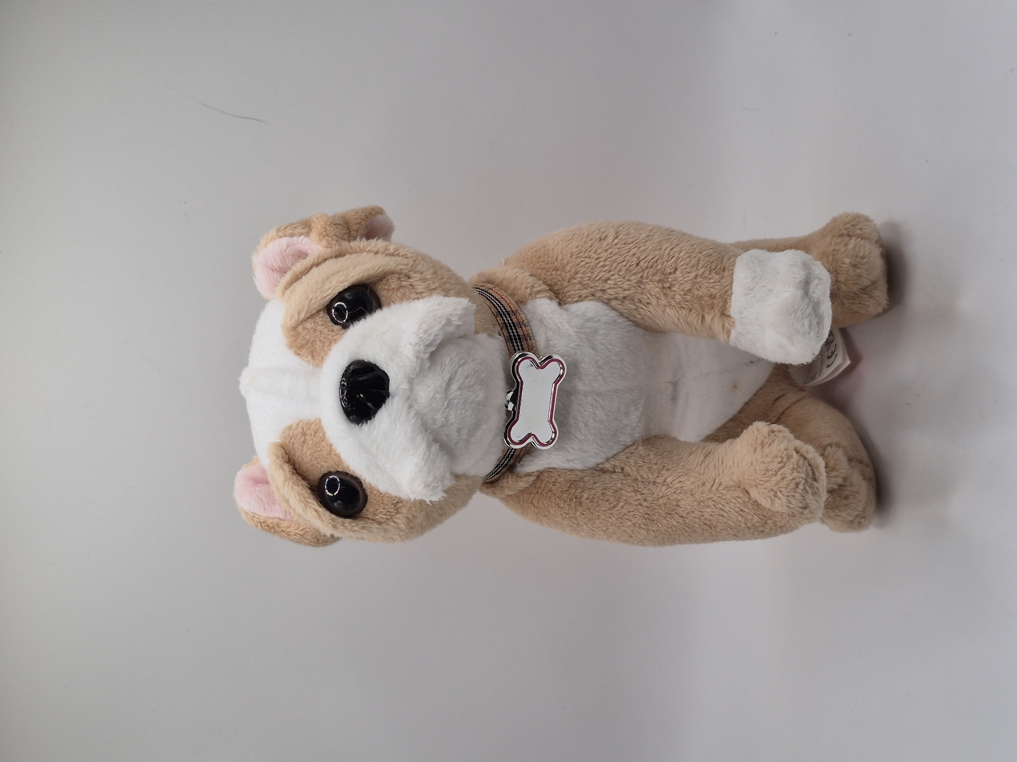 A stuffed bulldog sits facing forward against a plain, light gray background. The dog is tan and white with large, dark eyes, a black nose, and pink inner ears. It wears a plaid collar with a white, bone-shaped tag that has a pink outline and a blank space in the center. The dog's front paws are positioned in front of its body. There is no discernible indication of the city of Schwäbisch Gmünd, Germany, within the image.