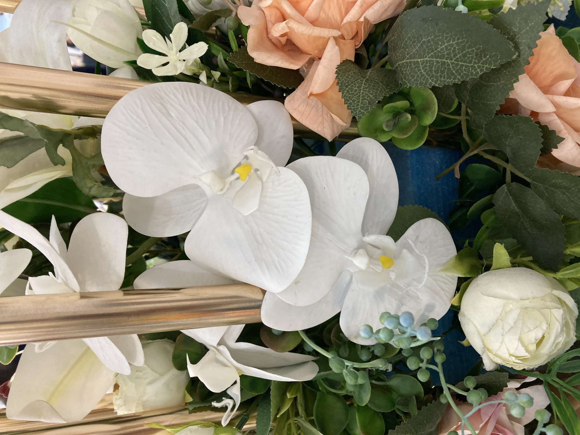 This is a close-up, top-down, eye-level shot of a floral arrangement. The arrangement is dominated by white orchids, with prominent yellow centers. Interspersed are peach-colored roses, a pale pink rose, and a small white rosebud. Green foliage, including succulents and small, bead-like clusters, fills the spaces between the flowers. Two vertical golden rods are visible, suggesting the arrangement is part of a larger display or structure. The background is a blurred blue surface. The image is a still life and does not depict any specific location within Tashkent, Uzbekistan, nor does it show any people or actions.