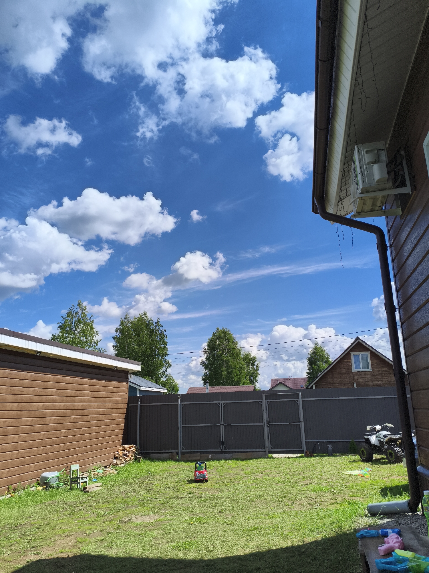 The image depicts a sunny outdoor scene at a residential property in Русский Вожой, Russia. The sky is a vibrant blue with scattered white cumulus clouds. The foreground features a green lawn. To the left, a brown, wooden-sided building with a white-trimmed roof is visible. Adjacent to this building, a small stack of firewood is present. Further into the yard, a dark gray metal fence runs horizontally across the middle of the frame. Beyond the fence, the roofs of other houses and trees can be seen. In the yard, there is a small red toy car and a white ATV parked near the fence on the right. The right side of the image is dominated by the side of another brown wooden house with a dark brown gutter system and an outdoor air conditioning unit mounted high on the wall. Several lengths of string lights are also visible hanging from the eaves of this house. The scene appears to be a backyard.