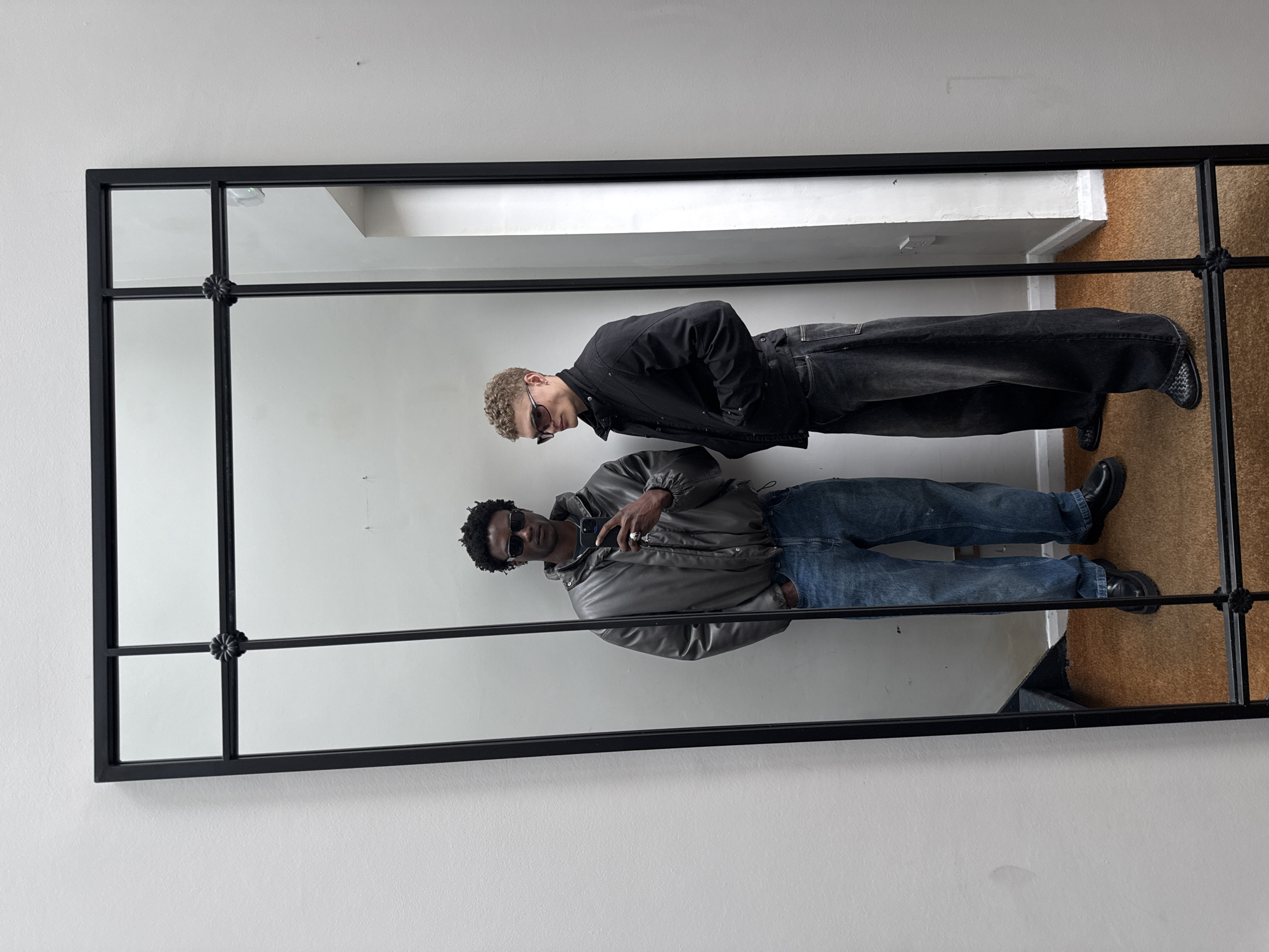 Two men are reflected in a large rectangular mirror with a black frame and decorative floral motifs at the intersections of the frame's internal divisions. The mirror is positioned against a white wall, and the floor visible in the reflection appears to be covered with a light brown material, possibly cork or carpet.
The first man, closer to the bottom of the frame, is wearing a grey puffer jacket, blue jeans, black shoes, and sunglasses. He holds a smartphone in his right hand, seemingly taking a picture of himself in the mirror.
The second man, located above the first in the reflection, is wearing a black jacket, dark jeans, sunglasses, and black shoes. His arms are crossed.
The setting appears to be indoors, possibly a residential space. The absence of landmarks prevents identification of a specific location in London. The lighting is ambient and even, illuminating the figures and the surroundings adequately.