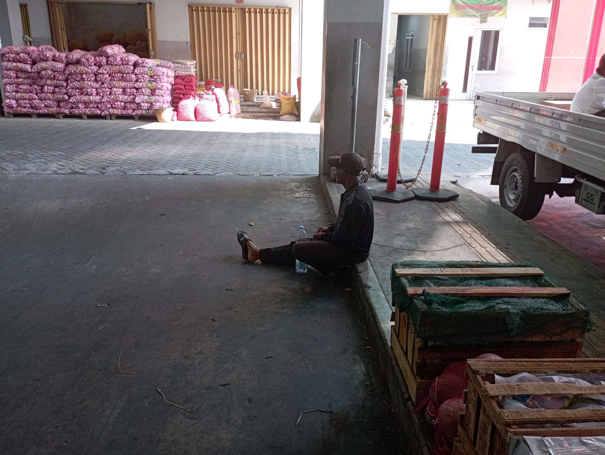 The image depicts a scene at the Ponorogo traditional market (Pasar Legi Ponorogo) in Ponorogo, Indonesia. In the foreground, a man wearing a dark shirt, dark pants, and a baseball cap is seated on the ground with his legs extended. He is facing to the right, away from the camera, and appears to be resting. A blue plastic water bottle is visible next to him.
Behind the man, large stacks of pink sacks, likely containing onions or similar produce, are neatly arranged on wooden pallets. Further back, more sacks of produce are visible, some in red netting and others in white. A truck bed, possibly a flatbed truck, is parked in the background on the right side of the image. Red and white traffic bollards with chains are also present, indicating a designated area or traffic control within the market. The ground is paved with interlocking bricks in the area where the produce is stacked, transitioning to a concrete surface where the man is seated. The environment suggests an active market setting during daylight hours, characterized by the storage and transport of goods.