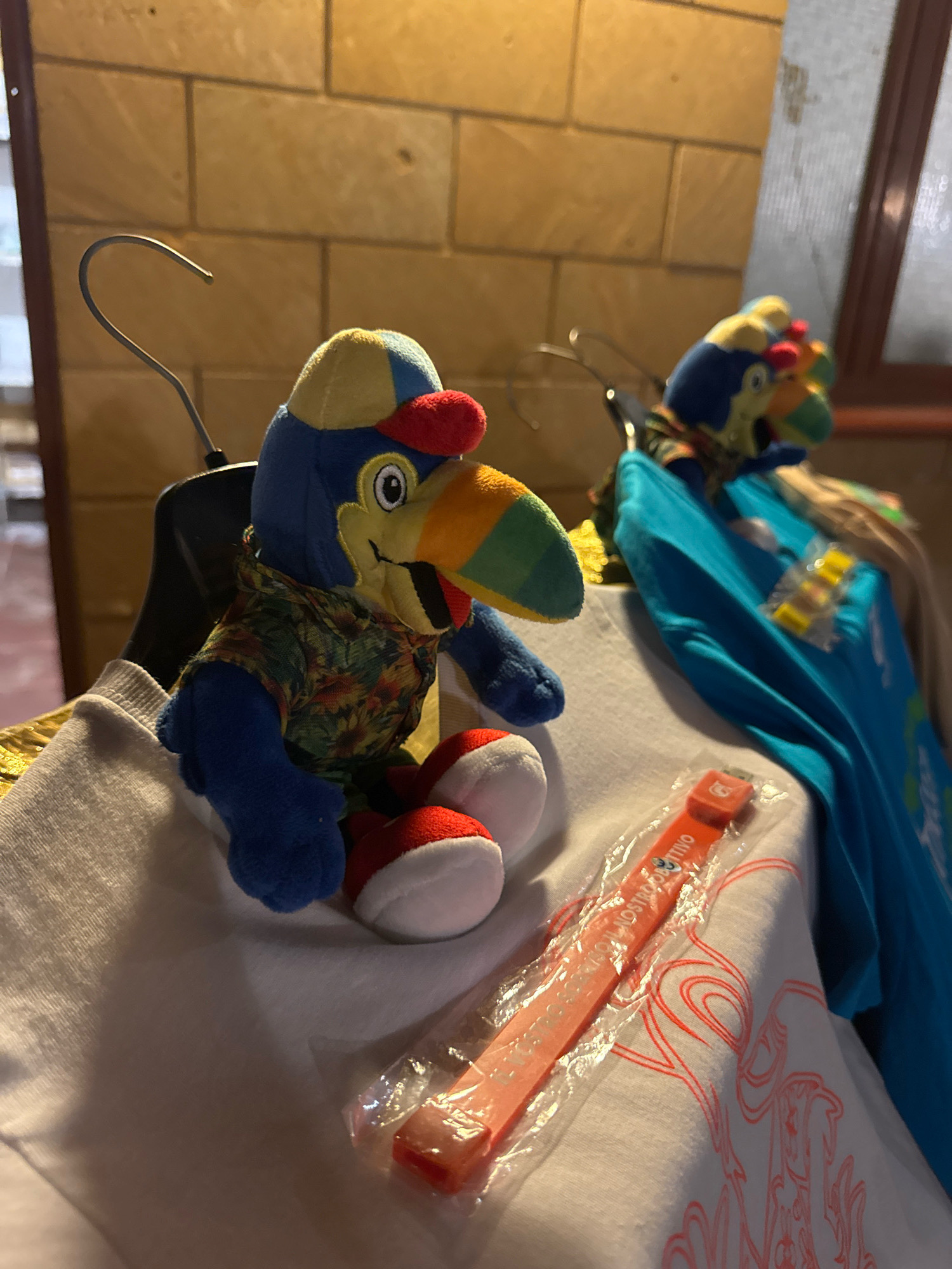 The image shows two plush toucans wearing hats and tropical shirts, one in the foreground and another in the background. The foreground toucan is sitting on a white garment, possibly a t-shirt, with a red and orange wristband in a plastic wrapper lying across it. The background toucan is partially obscured by a blue shirt hanging on a rack. The scene appears to be indoors, with a brick wall in the background. There are no people visible in the image. The lighting suggests it is evening or in a dimly lit area. The location is Isola delle Femmine, Italy.