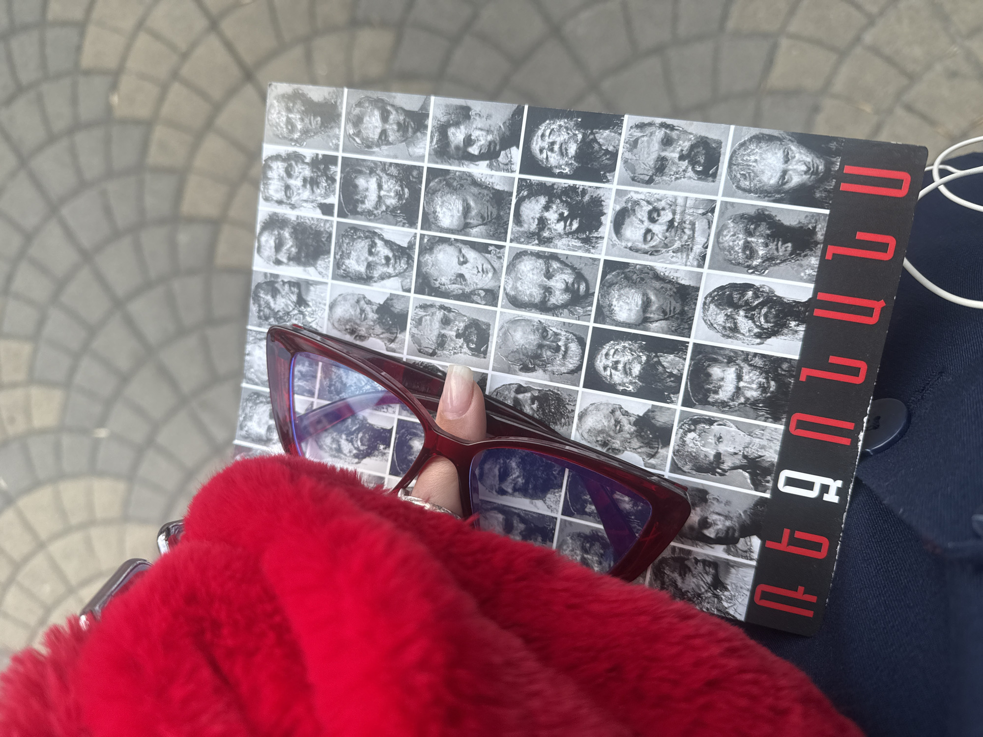 A hand holds a book with a black-and-white grid-like cover, overlaid by red-framed glasses, and rests on a blue-colored garment next to a tuft of red, fuzzy material. The book features numerous grayscale images that appear to be heads or faces arranged in a grid pattern. Red text runs vertically down the right side of the book cover. The glasses have a prominent angular frame and clear lenses. The hand has a manicured nail. A white earbud wire is also present. The background shows a cobblestone or paved surface.
There is no information about the location in the city of Yerevan, Armenia.