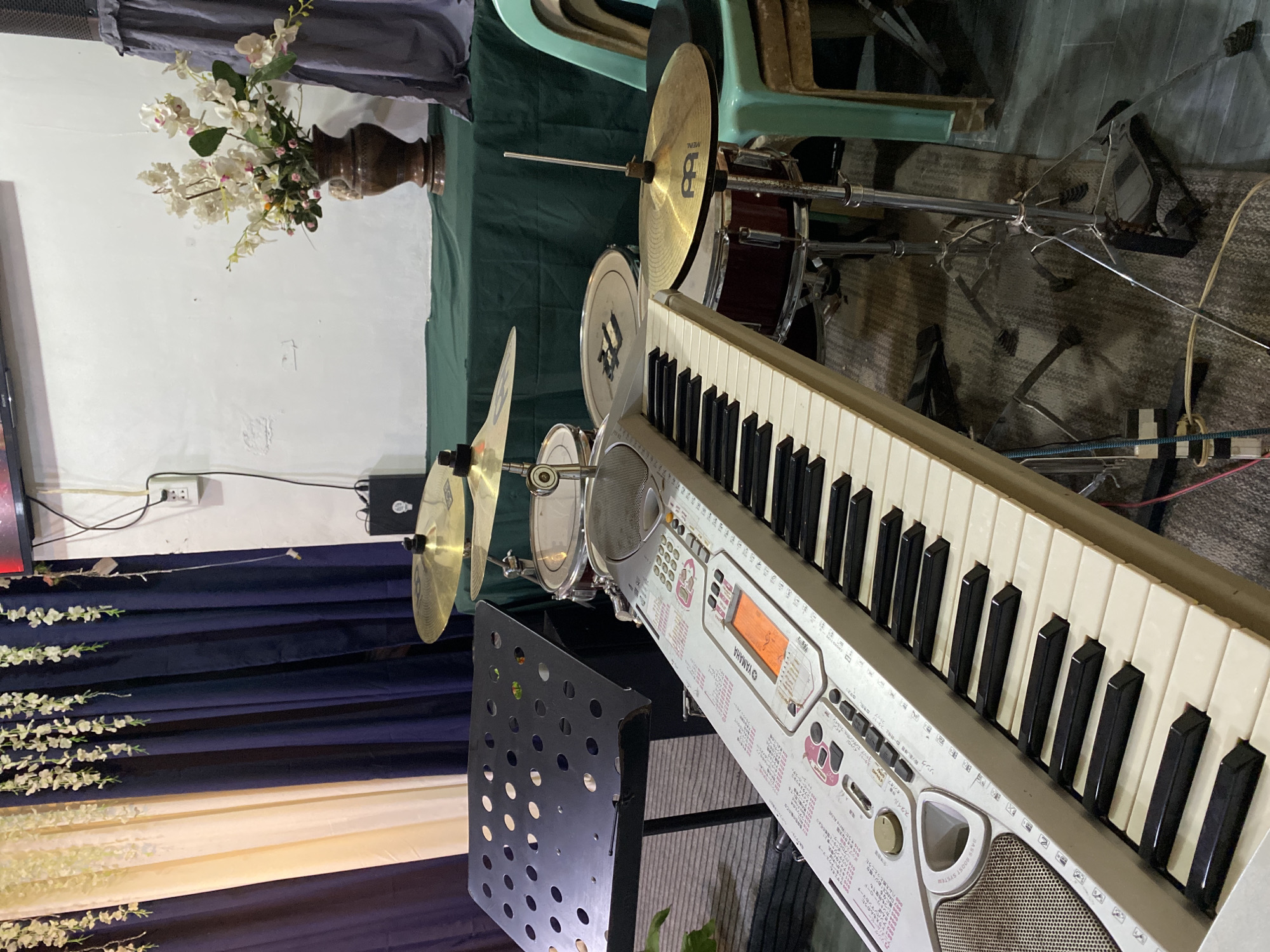 A Yamaha electronic keyboard is in the foreground, angled with its keys facing the viewer. To the right of the keyboard, a drum set is partially visible, including cymbals and a snare drum. A music stand is positioned to the left of the keyboard. In the background, there are draped curtains in navy blue and beige, and a vase with white flowers sits on a table covered with a green cloth. There are also some stacked chairs in the background. The image appears to be taken indoors, possibly in a church or event hall, given the decorations. The scene is set in Davao City, Philippines.