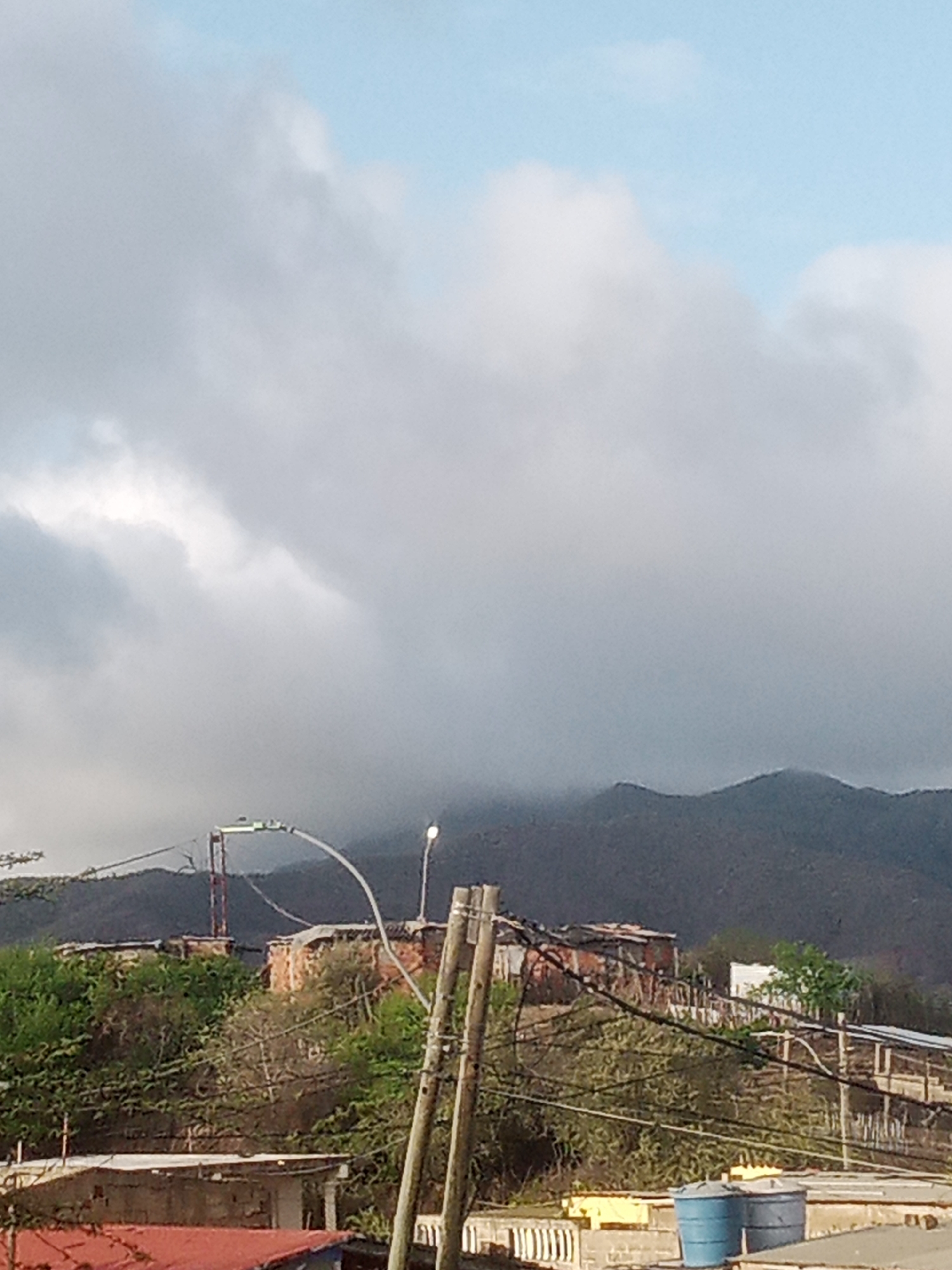 The image depicts a view of La Tagua, Venezuela, featuring a mountainous landscape partially obscured by clouds. In the foreground, a cluster of modest, mostly brick and corrugated metal structures are visible, nestled on a vegetated hillside. Electrical wires crisscross the scene, supported by wooden poles. A streetlamp can be seen illuminating the area. Two large blue water tanks are present in the lower right portion of the frame. The overall impression is that of a rural or semi-urban environment within a mountainous region.