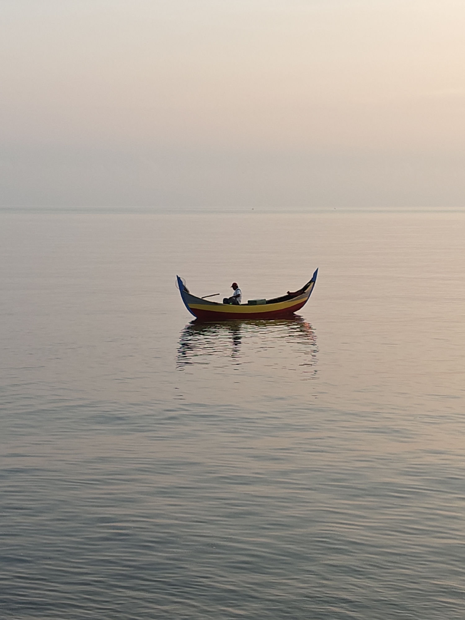 A lone figure rows a colorful, traditional boat on a calm body of water. The boat has a red hull, yellow interior, and blue trim. The person is wearing a white shirt and a red cap. The water has gentle ripples. The sky is a muted, hazy pink. The photograph was taken in Lhokseumawe, Indonesia.
