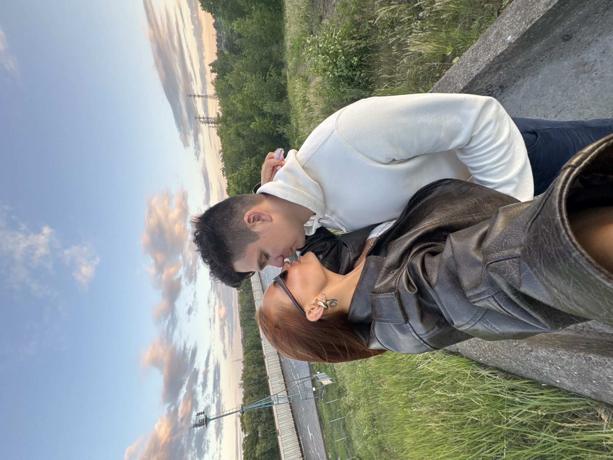 A couple is captured in a selfie kiss against the backdrop of a sunset in Chaikovsky, Russia. The woman, with auburn hair, wears a black leather jacket and sunglasses, her head tilted towards the man. She has large hoop earrings and a visible tattoo on her neck. The man, with dark hair, is wearing a cream-colored hoodie and dark pants. They are standing on a concrete pathway bordered by tall, green grass. In the background, a river flows past industrial structures and lush trees. Power lines and a tall tower with lights are visible on the horizon. The sky is painted with soft pink and orange clouds, indicating the end of the day. The overall setting appears to be an elevated path overlooking a scenic, semi-industrial area.