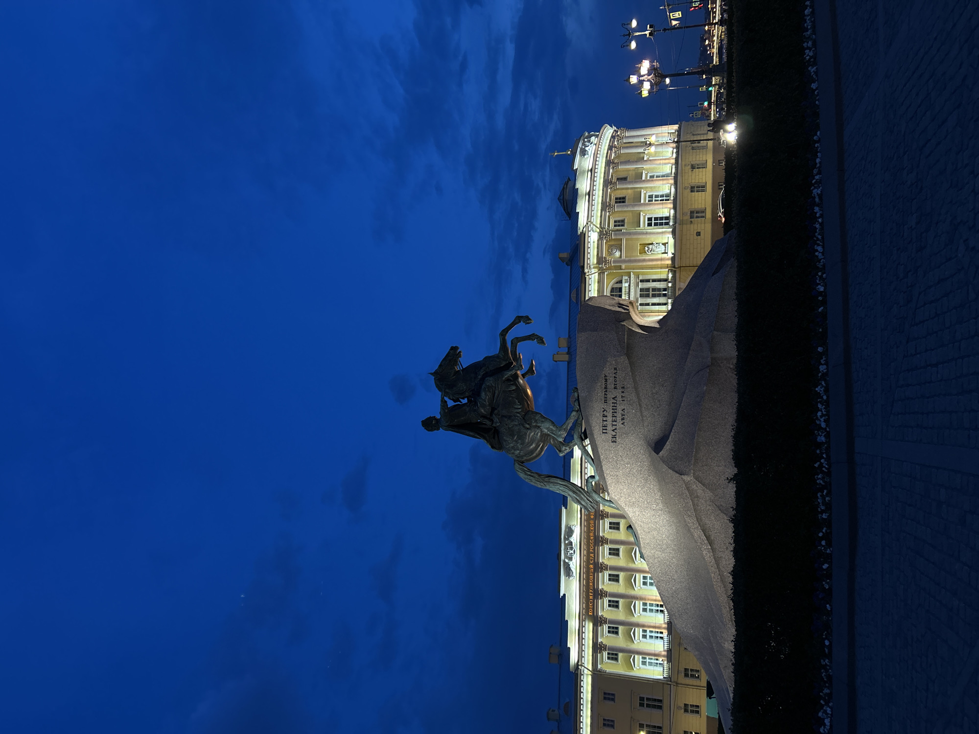 The image captures the Bronze Horseman statue in Saint Petersburg, Russia, at night. The statue depicts Peter the Great on horseback. The statue sits upon a large granite boulder, with inscriptions visible on its surface. Behind the statue is a brightly lit building with multiple windows and architectural details. The sky is a dark blue, indicating a nighttime setting. There are streetlights visible, adding to the artificial illumination. The foreground includes dark vegetation and what appears to be a paved area.