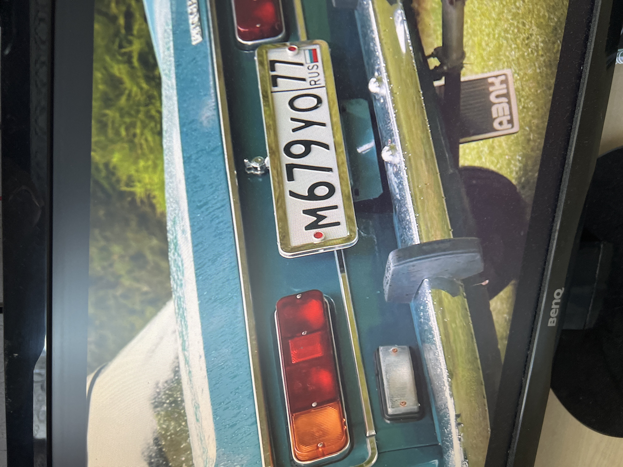 A rear view of a teal-colored vintage Soviet-era automobile, likely a Lada, is shown. The car is outdoors, on a grassy area, suggesting it might be parked on the side of a road or in a rural setting. The car's bumper and taillights are visible, and a license plate with Cyrillic characters and numbers is prominent. The license plate reads "M679YO 77 RUS," indicating it is registered in Russia, with "77" likely referring to the Moscow region. There is a mud flap with "AVAK" in Cyrillic on the lower right. The car appears wet, possibly from rain or dew. The scene is captured from a slightly elevated angle, looking down at the car. The background consists of green foliage. The image is a photograph of a screen displaying the car, as evidenced by the monitor's bezel at the top and bottom of the frame and the BenQ logo at the bottom. The specific location within the city of Kanash, Russia, cannot be determined from the visual information provided.