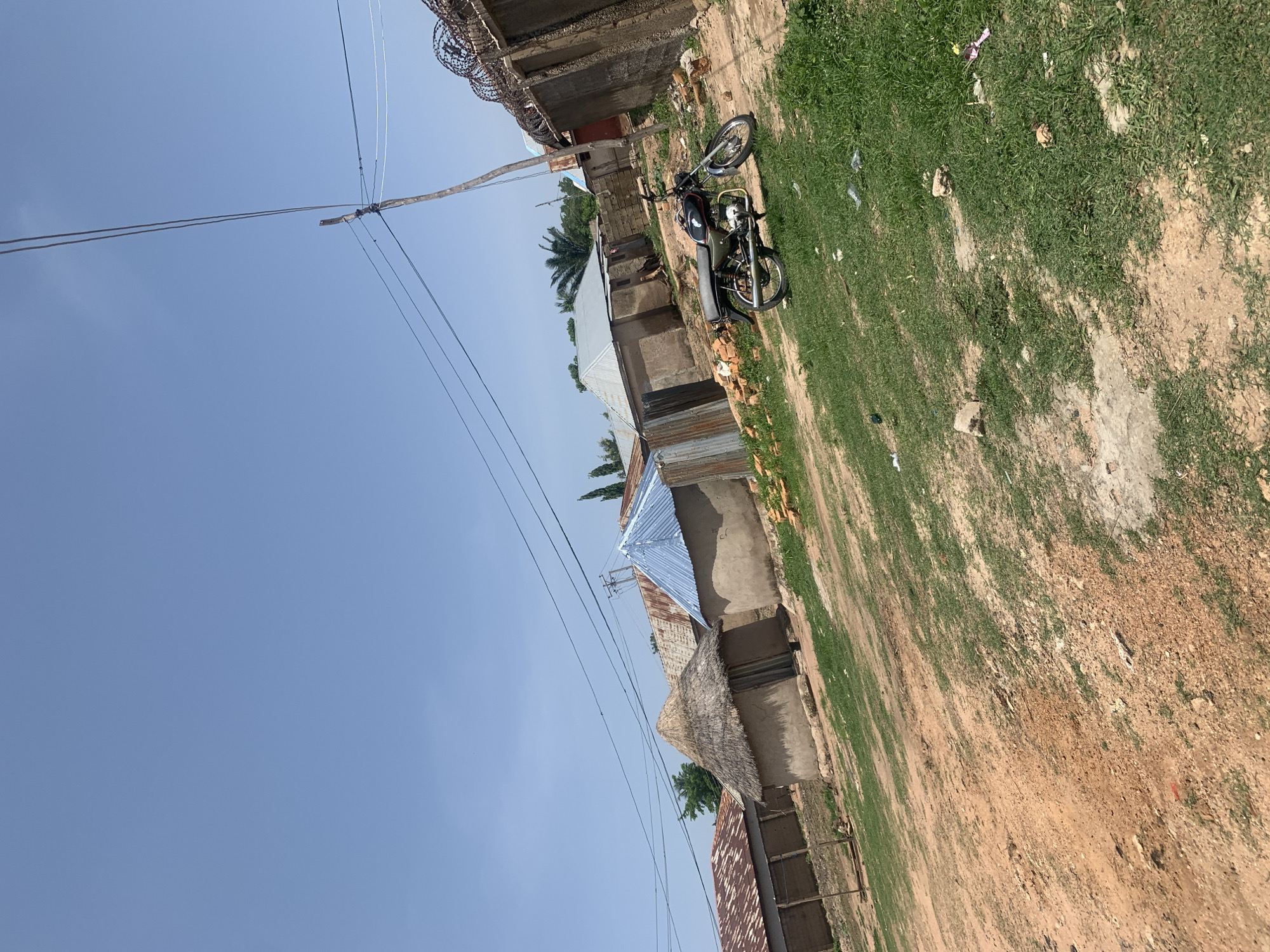 The image depicts a scene in Jauro Yinu, Nigeria, showing a cluster of buildings with various roofing materials, including thatch, corrugated metal, and what appears to be a blue metal roof. Some structures are made of mud or clay, characteristic of traditional housing in the region. In the foreground, a dirt path is visible, with patches of green grass and scattered debris. A black and silver motorcycle is parked on the right side of the path, leaning on its kickstand. A tall utility pole with overhead wires stands behind the motorcycle, and a fence topped with barbed wire runs along the right edge of the frame, bordering the property. The sky above is clear and blue. The overall impression is of a rural or semi-urban residential area in Nigeria under a bright, sunny sky.