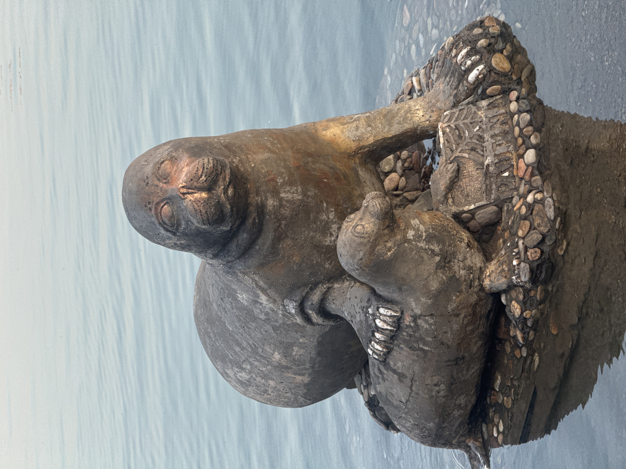 A bronze sculpture of two seals, one adult and one pup, sits on a rocky base in the water of the Angara River in Irkutsk, Russia. The adult seal is larger and is positioned behind the pup, with its flipper resting on the pup's back. Both seals are facing forward, with their heads slightly tilted upwards. The sculpture is weathered, with a patina of age and some green verdigris. The water is calm and reflects the sky, which is overcast. The rocky base is made up of various sizes and colors of stones, and some are submerged in the water. There is a small plaque embedded in the rocks at the base of the sculpture with some text on it, but it is not legible in the image. The scene is peaceful and serene.