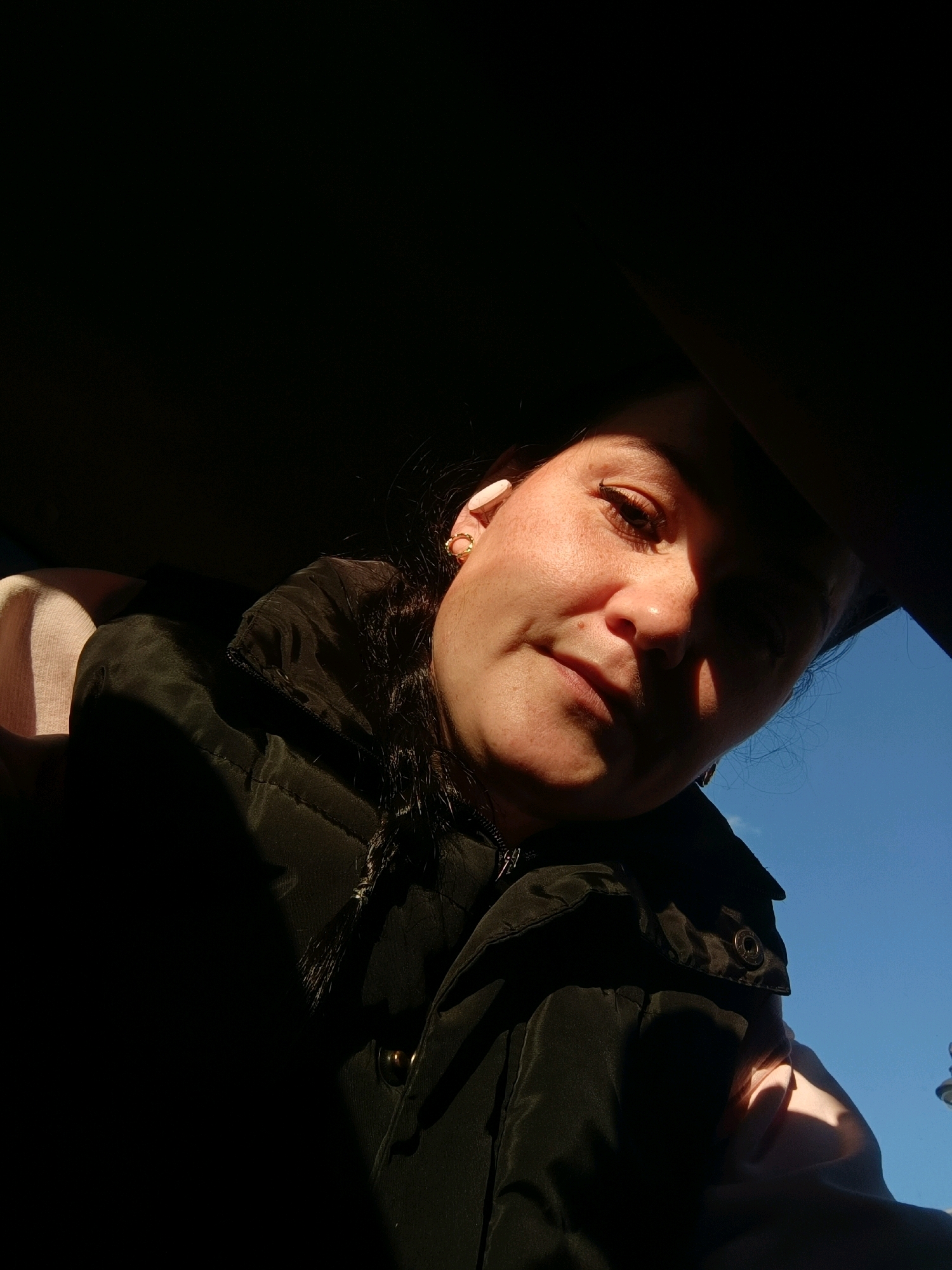 A woman is captured in a close-up, high-angle selfie taken from inside a vehicle, suggested by the dark interior framing the shot and the visible blue sky outside. The sunlight is casting dramatic shadows across her face, highlighting her features. She has dark hair styled loosely and is wearing a dark jacket. A gold earring is visible on her left ear. The location in Perm, Russia, is not discernible from the image itself.