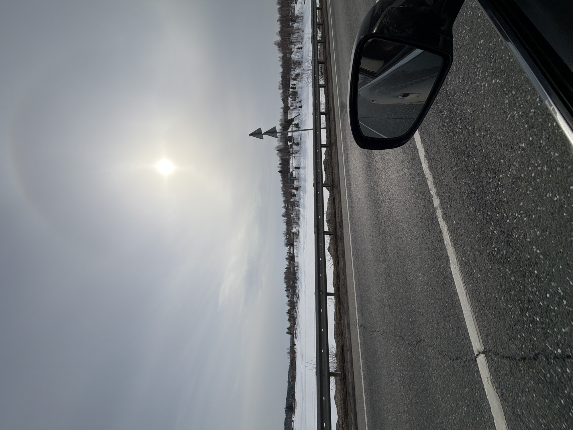 This image depicts a winter or early spring landscape in Ust'-Shalashnaya, Russia, viewed from inside a moving vehicle. The bright sun, positioned in the upper-middle right quadrant of the sky, exhibits a visible 22-degree halo. The sky is largely overcast or hazy light gray.
In the foreground, the asphalt road surface occupies the lower half of the frame, displaying dark grey aggregate and multiple visible cracks. A solid white line delineates the right edge of the visible roadway. A black vehicle side mirror is prominent in the lower right quadrant, reflecting a portion of the vehicle's light-colored bodywork and the road behind. The dark frame of a car window is visible immediately below the mirror.
A silver metallic guardrail runs horizontally across the midground, separating the road from the adjacent terrain. Beyond the guardrail, the ground is covered extensively in snow, indicating cold conditions. Sparse, dark, deciduous vegetation, primarily bare bushes and small trees, protrudes from the snow. Two dark, triangular road signs are vertically stacked on a single pole, situated to the right of the frame, between the guardrail and the snow-covered field.
In the background, a distinct tree line consisting of bare deciduous and some coniferous trees marks the horizon. Behind this tree line, scattered low-rise buildings are visible, suggesting a rural or suburban settlement. No individuals, explicit actions, or direct interactions are visible within the frame.