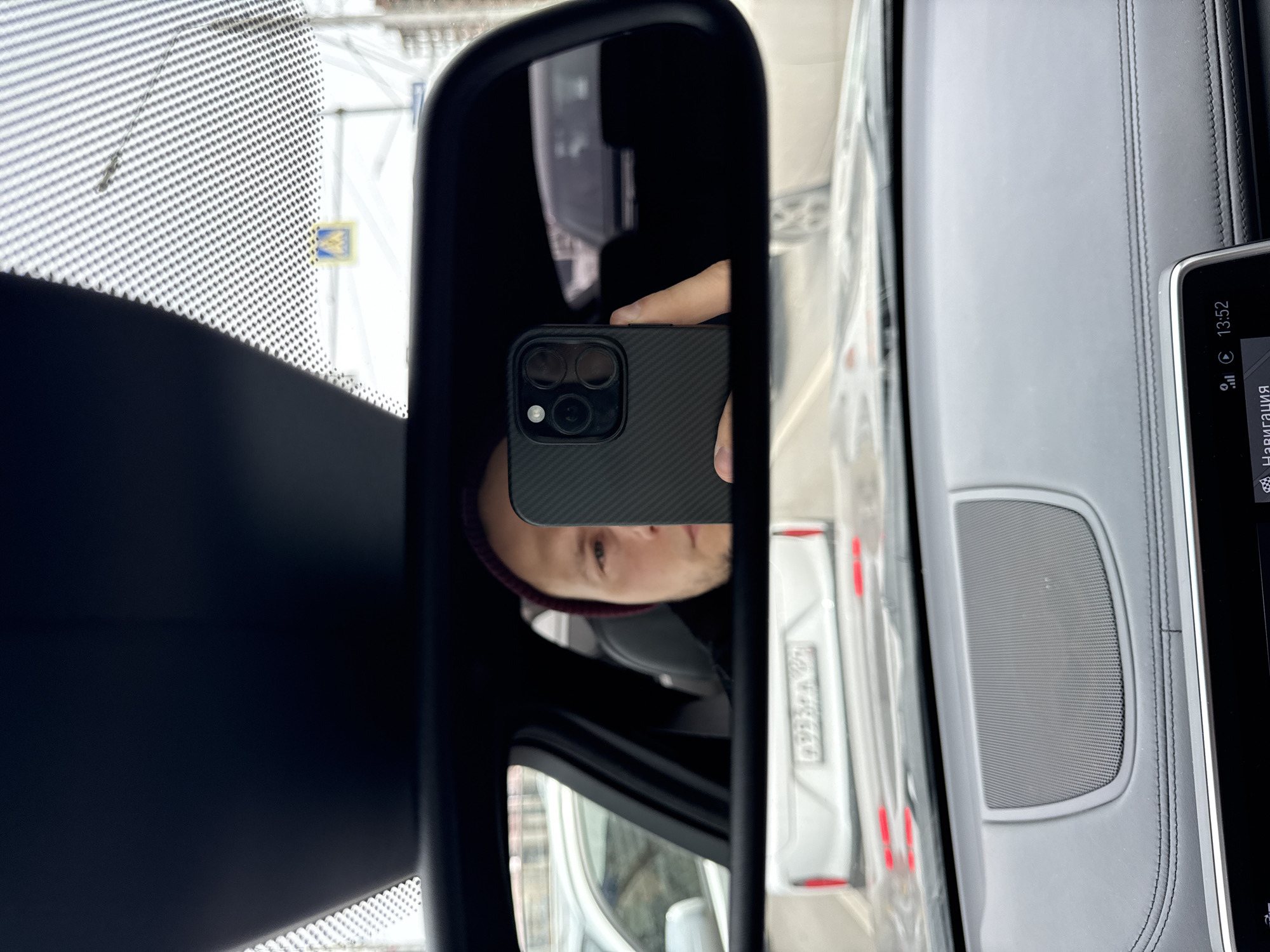 A rearview mirror reflects a person holding a smartphone. The mirror's reflection shows the person's face, part of their head, and a white car in the background. The car's license plate is visible but not entirely legible. Outside the car, a pedestrian crossing sign is present. The smartphone's camera section is visible. The photo appears to be taken from inside the car with a view of the dashboard. The time on the car's dashboard is 13:52. The presence of Cyrillic characters on the dashboard suggests the photo was taken in Russia. The license plate format and pedestrian crossing sign are also consistent with Russia. This could be Krasnoyarsk due to the architectural style reflected.