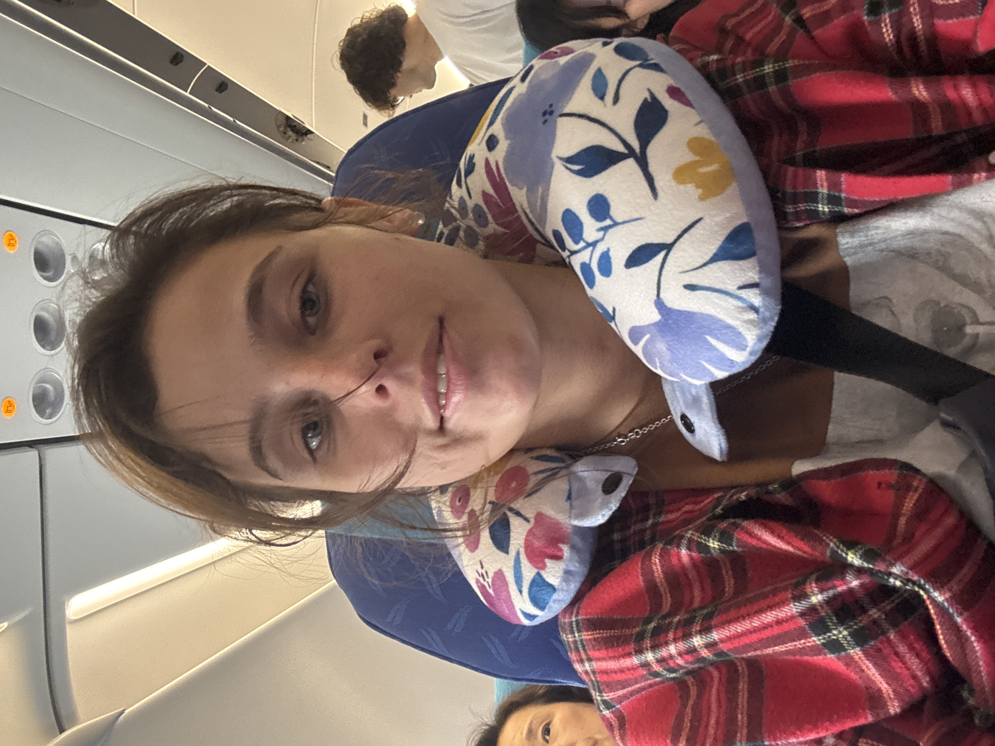 A woman is taking a selfie on an airplane, wearing a red and black plaid shirt and a floral patterned neck pillow. She has brown hair and is smiling faintly. To her left, a woman of Asian descent looks out of frame, and to her right, a man with curly brown hair is visible from behind, wearing a white t-shirt. The environment appears to be the interior of an airplane cabin, with overhead compartments and lighting visible. The image does not provide any information about the city of Busan, South Korea.