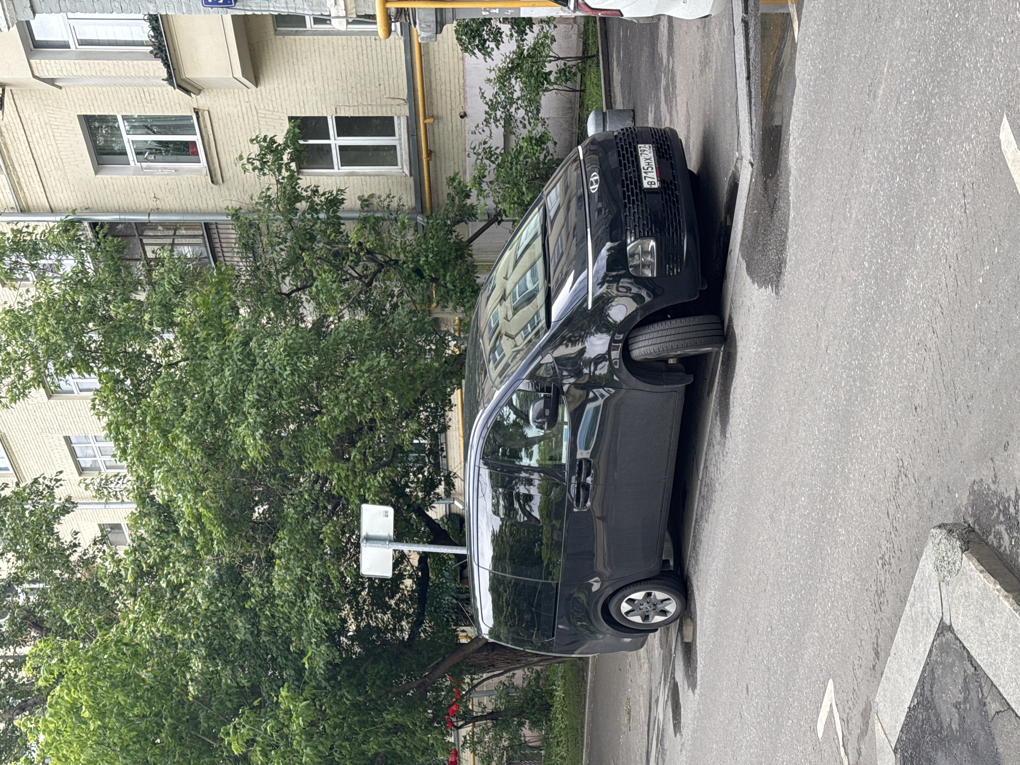 The image depicts a black Hyundai Staria parked on the side of a street in Moscow, Russia. The license plate number is B715HX797. A light yellow brick building with several windows is visible in the background. Green trees dominate the left side of the frame. The street is paved with asphalt, and a curb is visible on the right.