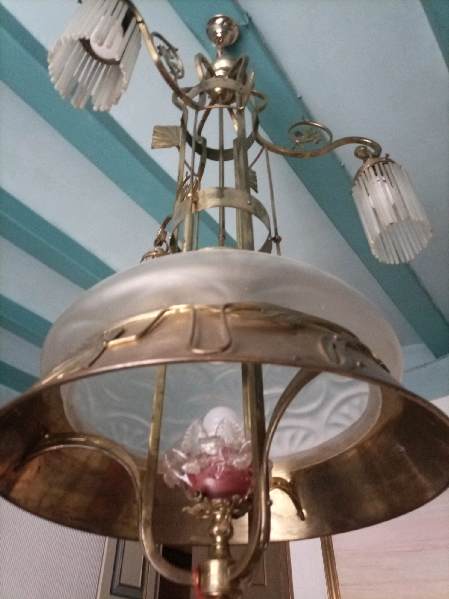A brass chandelier with a frosted glass shade hangs from a ceiling with blue and white striped wallpaper. The chandelier has several arms, each holding a cylindrical glass shade with vertical ridges. At the center of the lower part of the chandelier, a decorative element resembling a flower made of frosted glass with pinkish hues is visible. The overall style of the chandelier appears to be art nouveau or art deco. The image is taken from a low angle, looking upwards at the chandelier. The background indicates an interior setting, likely a home or public space in Tarragona, Spain.