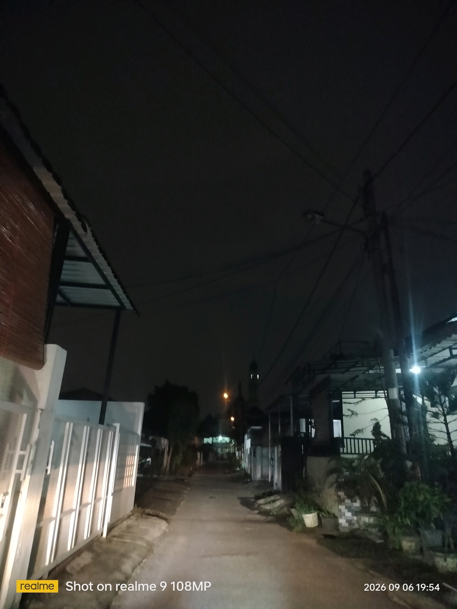 A nighttime photograph captures a dimly lit residential street in the City of Medan, Indonesia. The scene is dominated by dark tones, with streetlights and lights from houses providing the primary illumination. On the left side of the street, a white fence is illuminated by bright, vertical lights, suggesting they are embedded in or shining onto the fence. Adjacent to the fence is a building with a brown, slatted window covering and a dark, sloped roof supported by visible beams.
The street itself is paved and curves slightly into the distance, disappearing into darkness. On the right side of the street, houses with covered porches and windows are visible. One house has a dark railing on its porch. Several potted plants are placed outside the houses. In the far distance, a minaret of a mosque is faintly visible, silhouetted against the night sky. Overhead, a tangle of power lines stretches across the frame, attached to a utility pole that also has a street light. The sky is dark and overcast, with no stars visible. The bottom left corner of the image contains a "realme Shot on realme 9 108MP" watermark, and the bottom right corner displays the date and time "2026 09 06 19:54".