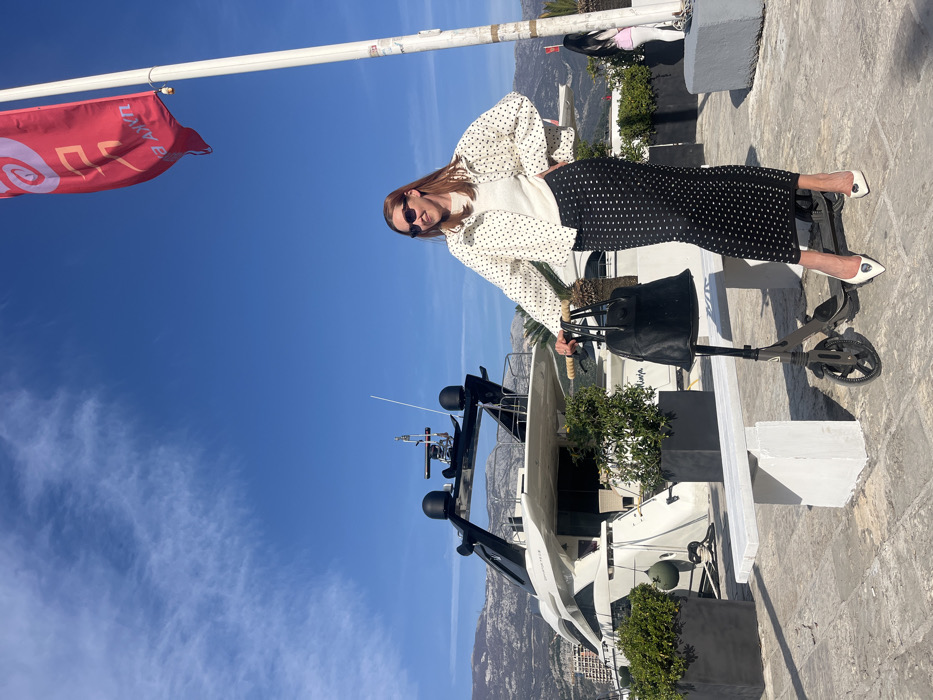 An adult female is standing on a paved promenade within a marina in Budva, Montenegro, under a clear blue sky with sparse clouds. She is positioned slightly to the right of the image center, facing the camera with a slight smile. Her hair is reddish-brown, and she wears dark sunglasses. Her attire consists of a white long-sleeved top beneath a white jacket patterned with black polka dots, a long black skirt also adorned with white polka dots, and white pointed-toe high-heeled shoes featuring a metallic emblem on the vamp. Her left hand is resting on the handlebar of a dark grey electric scooter parked to her immediate left. A black, possibly fur, handbag with light brown wooden handles is suspended from the scooter's handlebar. Her right hand is placed on her hip.
In the background, a large, multi-deck white motor yacht with black structural accents and radar equipment is docked. Behind the yacht, distant, bare mountains are visible. To the far right, a tall white flagpole flies a red flag displaying white Cyrillic text, possibly a logo or advertisement. The promenade is constructed from light-colored pavers and features two white concrete benches and multiple dark square planters containing green foliage. The scene is illuminated by strong sunlight, indicating an outdoor, daytime setting.