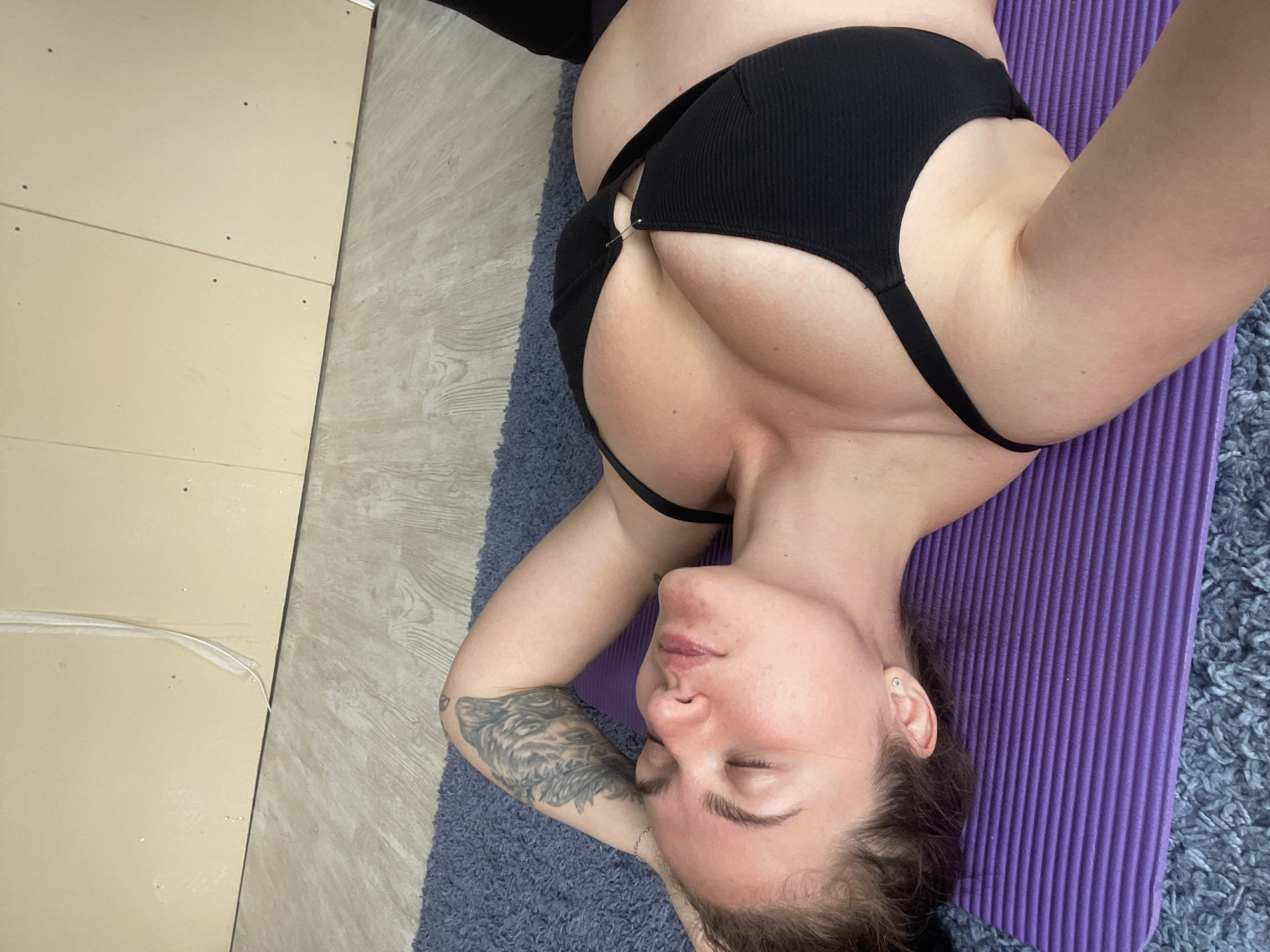 A woman lies supine on a purple yoga mat atop a blue shag rug. She wears a black ribbed sports bra. Her arms are bent and positioned behind her head. Her eyes are closed. A tattoo is visible on her left forearm. The backdrop includes beige wall panels and a light-colored floor. The location is a room in an unidentified building, not identifiable as being in the city of Мосметаллоконструкция Онт, Russia.
