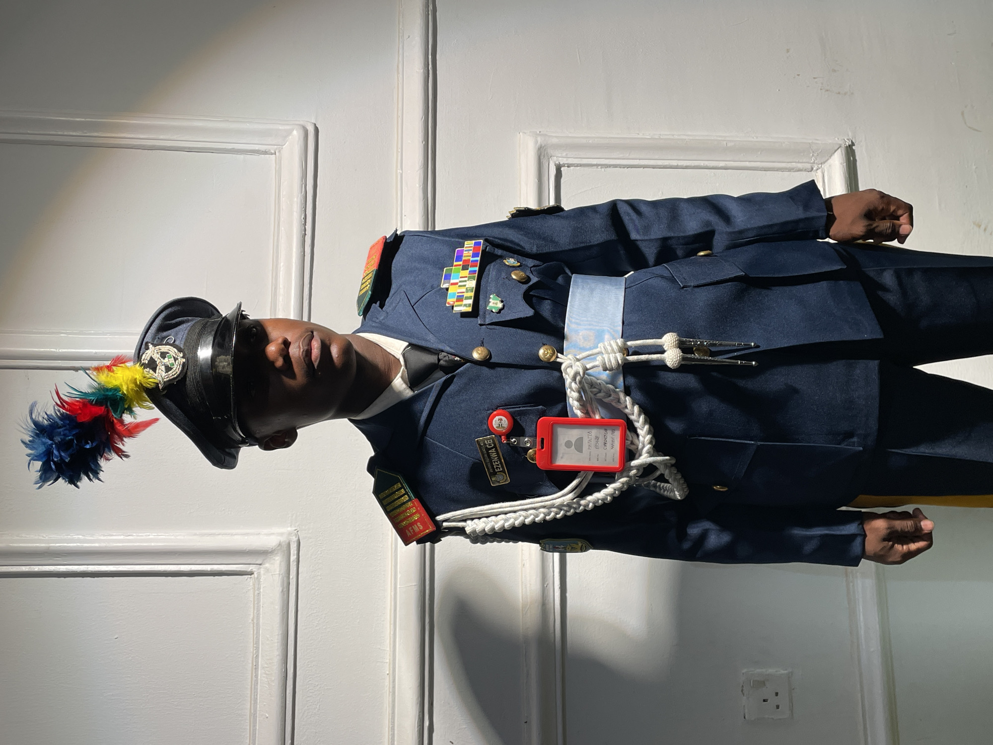 A medium indoor shot captures a person in uniform standing against a white paneled wall. The person, likely a member of the armed forces, is wearing a navy blue uniform jacket with various insignia and medals. They are also wearing a hat with a colorful plume. A lanyard and an ID card are visible on the uniform. The wall behind them has a paneled design, and an electrical outlet is visible near the bottom right corner of the frame. The city location, Barakin Akawo, Nigeria, is provided for contextual reference.