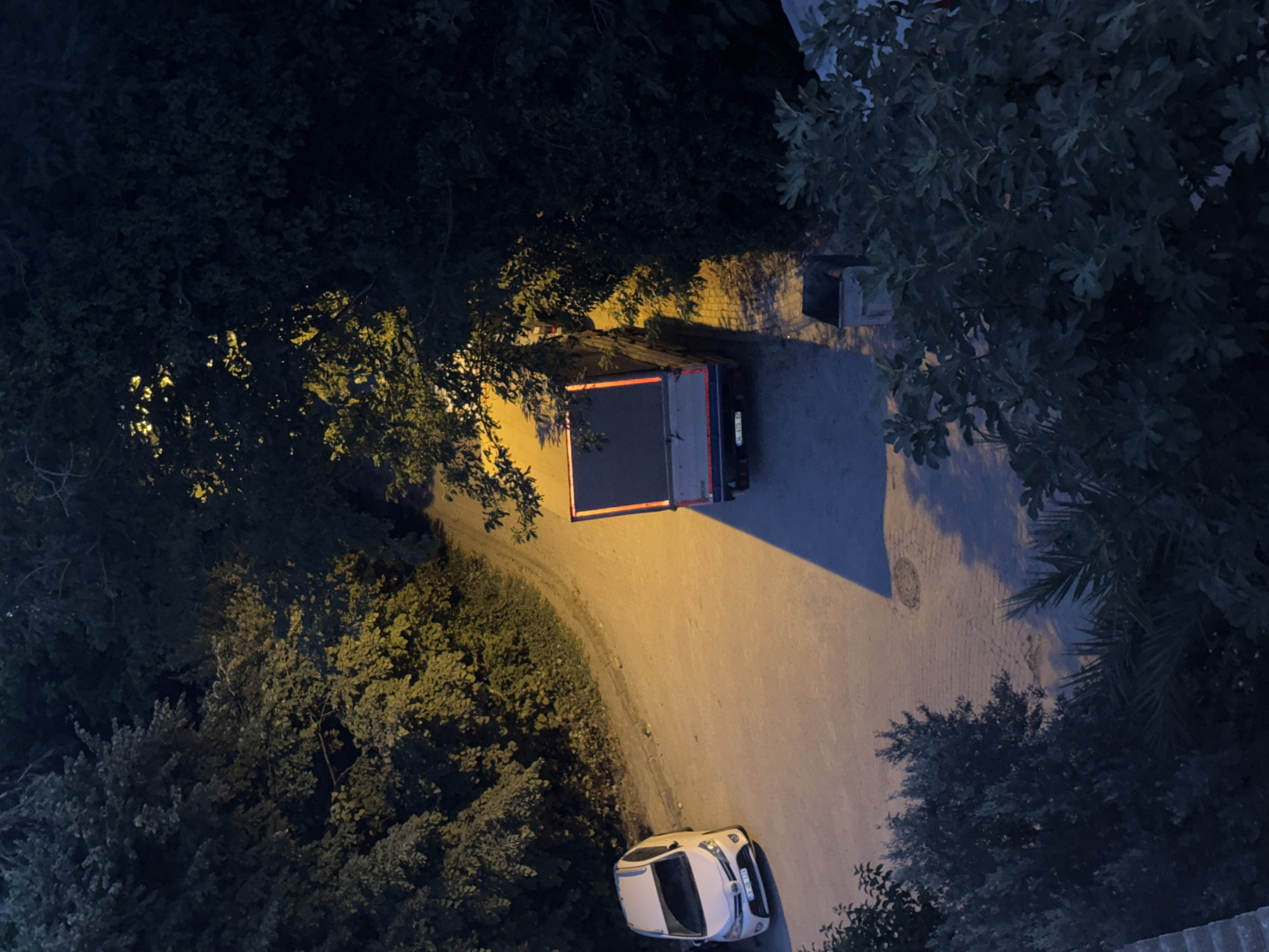 In the city of Gungoren, Turkey, an overhead view captures a dimly lit street scene at dusk or night. The street, partially illuminated by a warm yellow light, curves to the left. A white car is parked on the left side of the road, facing upwards. To the right of the white car, a flatbed truck is parked, its back end facing the viewer. The truck is dark with red taillights visible. The scene is framed by dense, dark green foliage on both sides of the street. The ground appears to be paved.