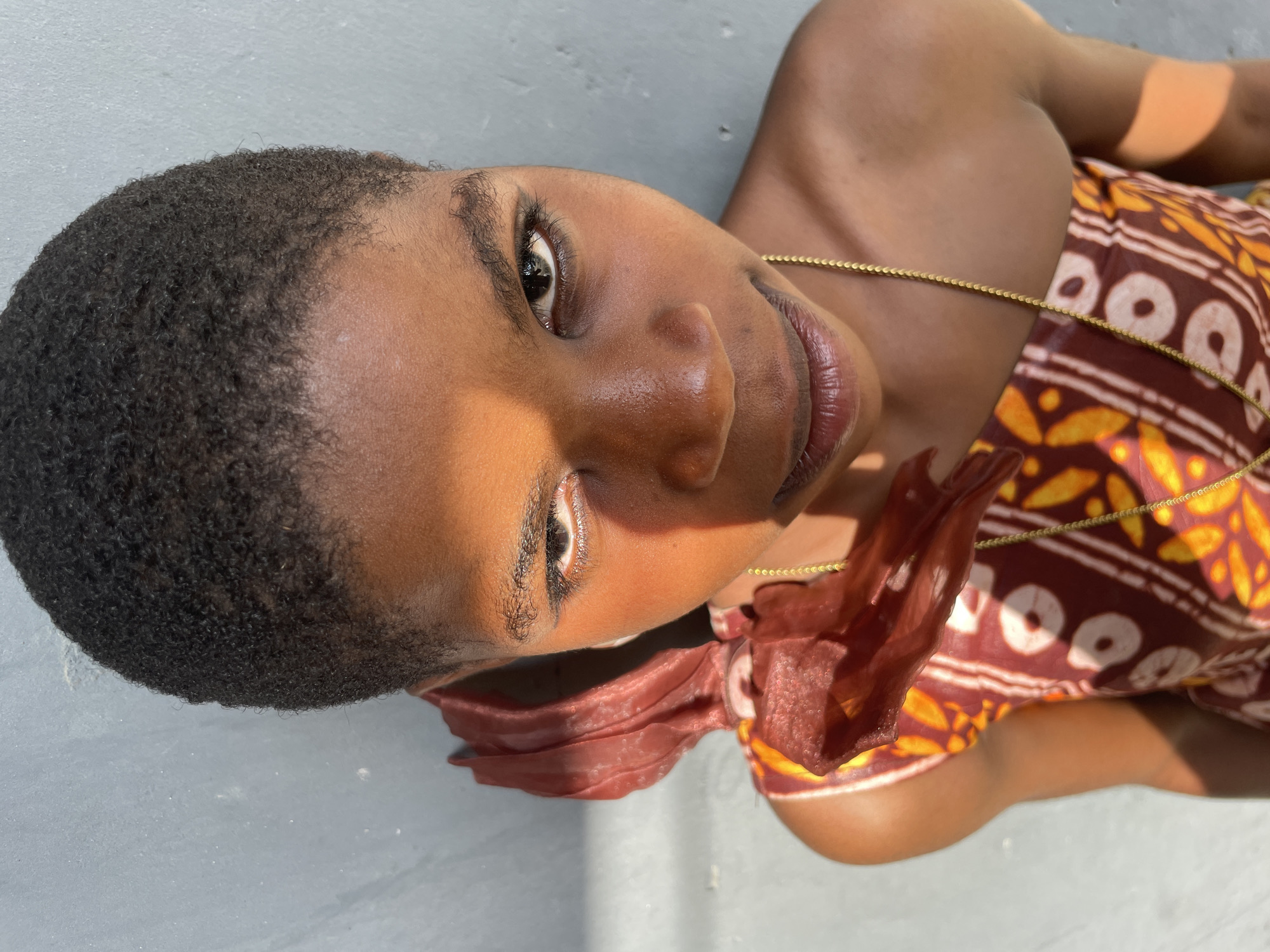 This close-up portrait, likely taken outdoors in Badagry, Nigeria, features a person with dark skin and short, tightly-curled dark hair. The subject is positioned in a striking manner, lying down and looking upwards and slightly to the right from the viewer's perspective, with their head tilted. The photograph is presented sideways, suggesting an artistic framing or the subject resting on a surface.
The person is wearing a vibrant garment with a distinctive pattern in shades of brown, white, and yellow-orange, adorned with circular and abstract leaf-like motifs. A thin, gold-colored chain is draped around their neck and across their chest. A piece of reddish-brown, possibly sheer or crinkled, fabric is visible near their right shoulder, adding texture to their attire.
The lighting is a notable element, with strong, direct sunlight casting dynamic shadows across the subject's face. The right side of their face (from the viewer's viewpoint), including their forehead and eye, is brightly illuminated, while the left side is partially shaded, creating a stark contrast and highlighting facial contours. Their dark eyes are open, looking directly towards the camera with a serene or contemplative expression.
The background is a plain, light gray surface, possibly concrete or a wall, against which the person is resting. The strong sunlight also casts shadows on this surface. There is no visible text in the image. The composition draws attention to the subject's features, the texture of their hair, and the intricate pattern of their clothing.
