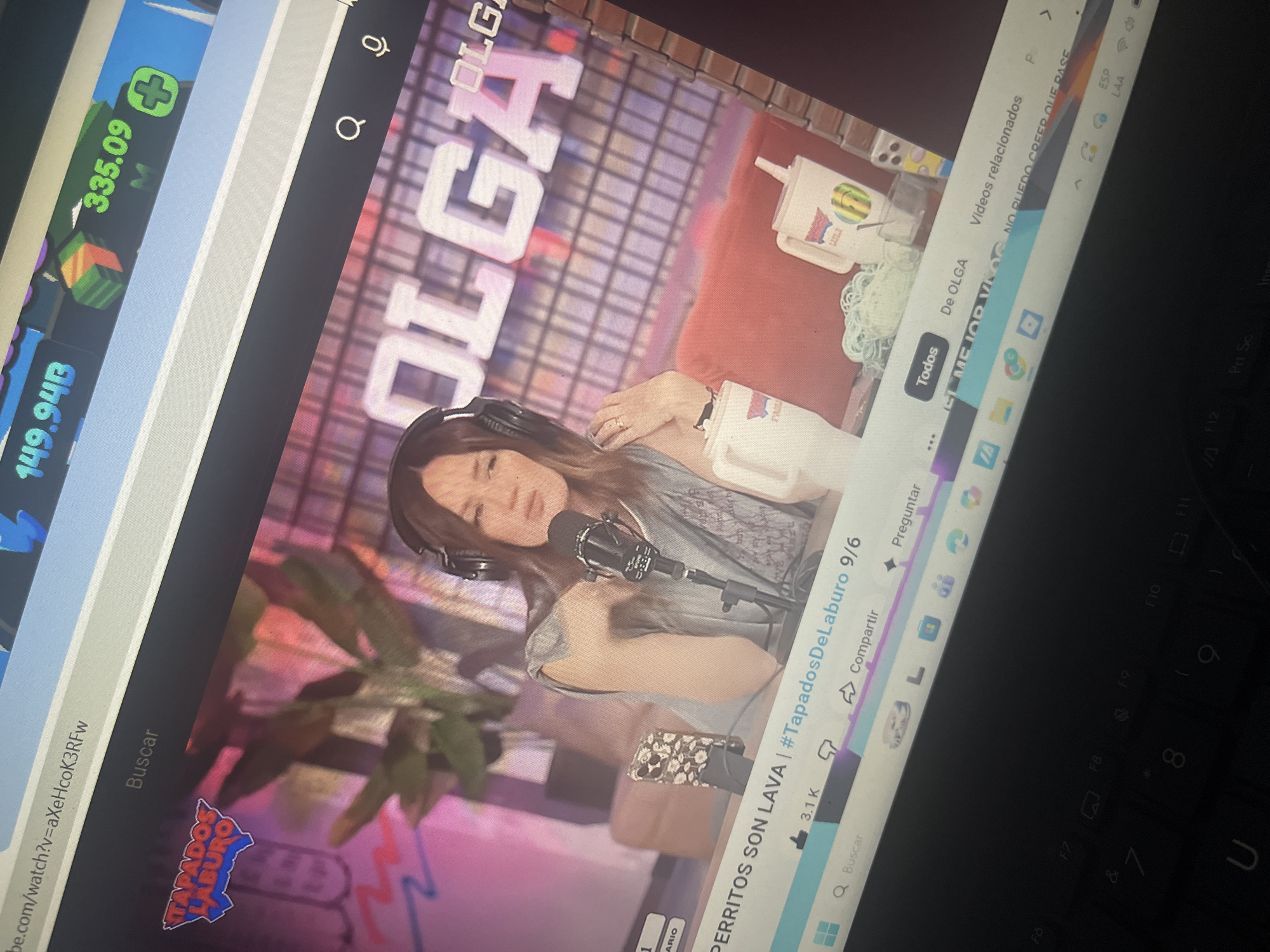 The image shows a laptop screen displaying a YouTube video. The video features a woman with headphones sitting in front of a microphone, with the word "OLGA" prominently displayed in large letters behind her. The video title appears to be "PERRITOS SON LAVA | #TapadosDeLaburo 9/6". Below the video player, there are typical YouTube interface elements like a "Like" button with "3.1K" indicating likes, a "Dislike" button, a "Share" button, and other icons. The overall setting appears to be a studio or a room set up for recording. The keyboard of the laptop is visible in the foreground, suggesting the image was taken by someone using the laptop. There is no indication in the image that it is located in Santa Ana, Venezuela.