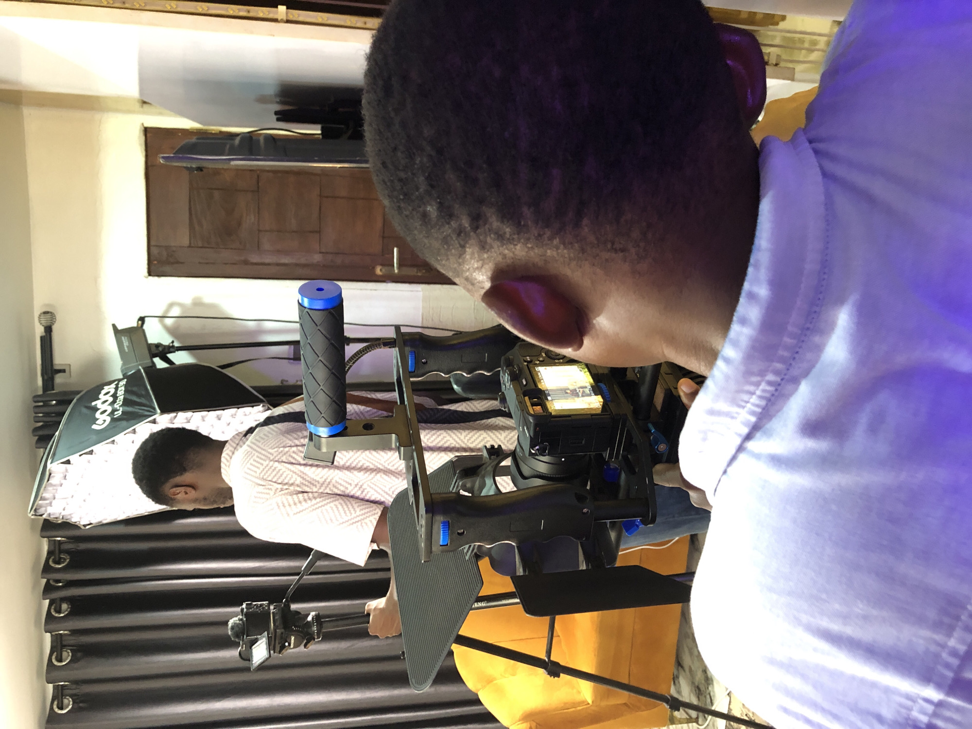 The original image is oriented vertically and depicts an indoor setting, likely a film or photography studio, with two men engaged in a production activity.
In the foreground, seen from behind and from a high angle, is a man with short, dark hair, wearing a light-colored collared shirt that has a purple tint, possibly from artificial lighting. He is looking down at a complex camera rig, which he appears to be operating or setting up. The rig features multiple handles, one with a prominent blue cap and textured black grip, and holds a professional-grade camera with its screen displaying an image or video feed.
Further in the background and to the left, another man is partially visible, also seen from behind. He is wearing a light-colored shirt with a subtle pattern (possibly checkered or striped) and is standing near a large softbox light. The softbox, bearing the brand name "Godox," is mounted on a stand and illuminates the scene.
Other visible objects include dark, heavy curtains running horizontally across the middle of the frame, a wooden panel or door behind the second man, and a yellow chair or cushion. Below the second man, a tripod with what appears to be another camera or monitoring device is visible. The lighting seems to be artificial and controlled, creating a professional setup. There is also an electrical strip or molding visible along the top edge of the wall.