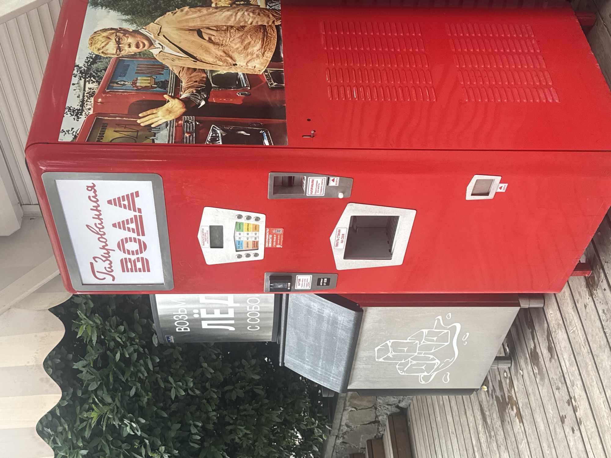 A red soda vending machine stands next to a metal ice dispenser on a wooden deck, with a lush green hedge in the background. The vending machine features a sign in Russian that reads "Газированная Вода" (Carbonated Water) and an image of a young man in a brown jacket and glasses. The ice dispenser has a graphic of ice cubes. The scene appears to be outdoors, possibly in a public area like a park or promenade in Yalta, Ukraine. The wooden deck is wet in places, suggesting recent rain or spillage.