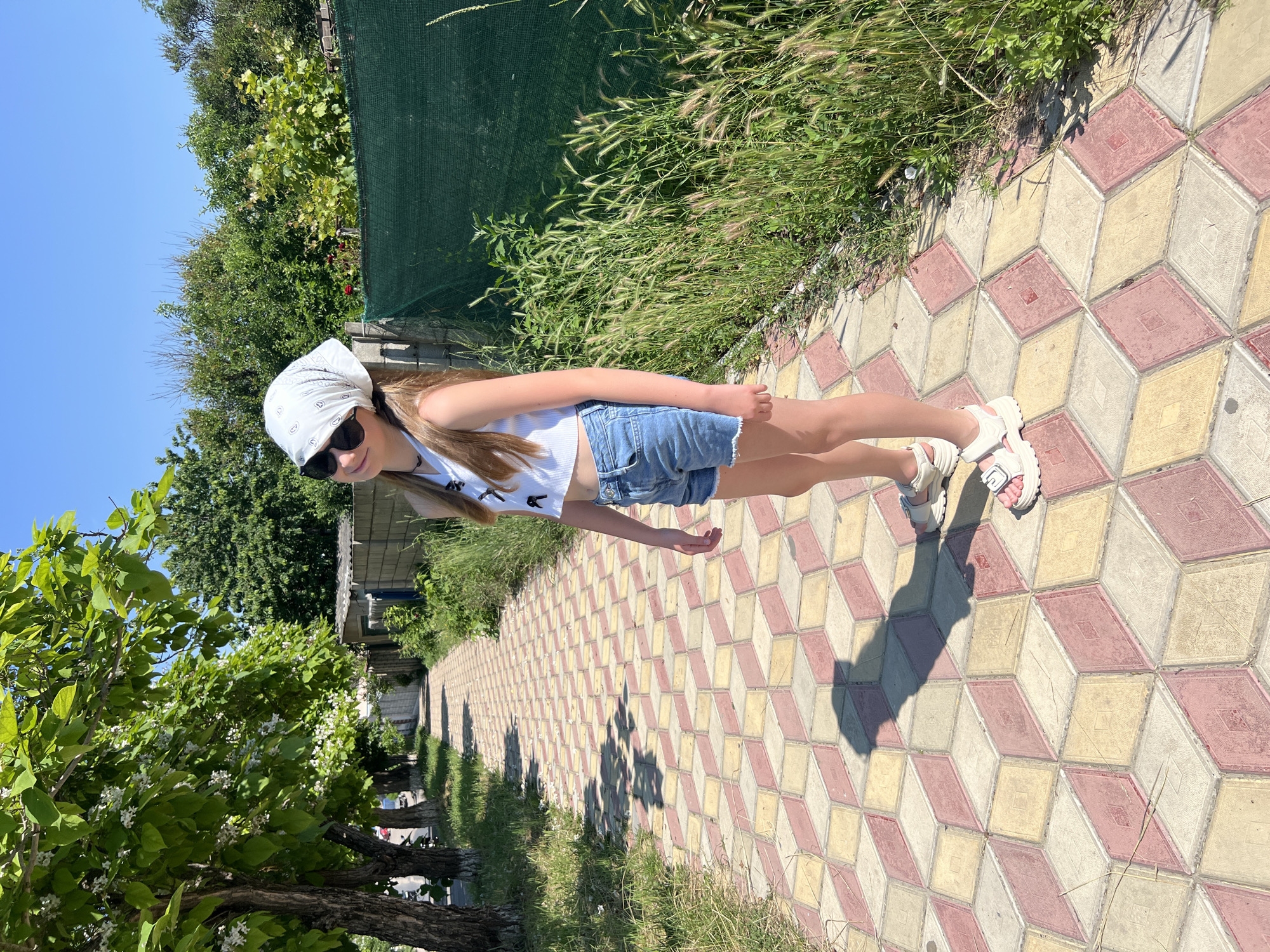 A young Caucasian girl with long blonde hair, wearing a white bandana, sunglasses, a white crop top, and denim shorts, stands on a patterned sidewalk in Dubăsari town council, Moldova. She is looking towards the camera with a slight smile. The sidewalk is made of red, yellow, and gray diamond-shaped pavers arranged in a 3D optical illusion pattern. To the left of the sidewalk, there are trees with green leaves and white flowers, and to the right, there is tall green grass and a dark green fence. The sky is clear and blue, indicating a sunny day. The girl is casting a shadow on the sidewalk.