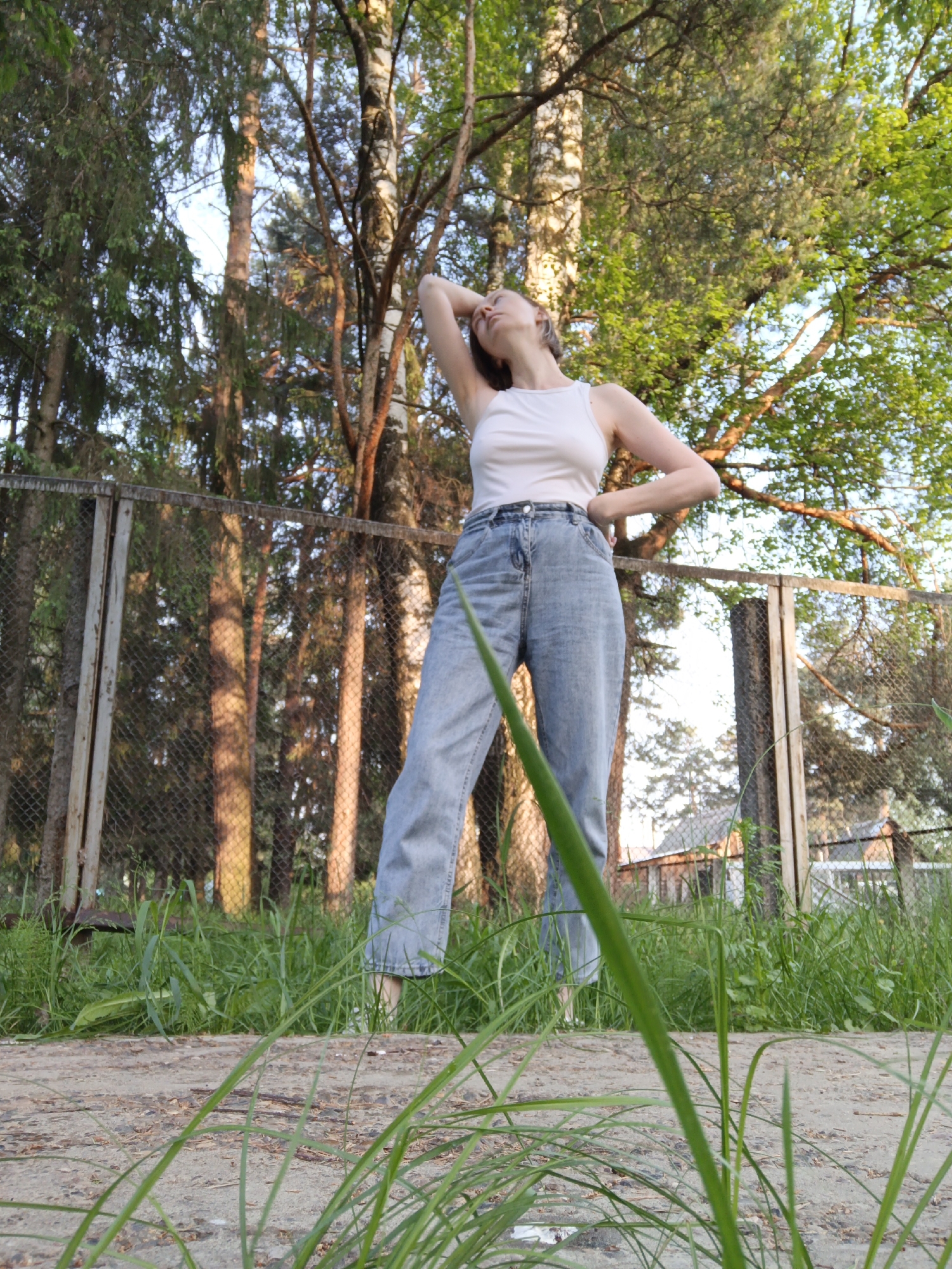 A full-length photo, taken at ground level, captures a young woman in a white tank top and light-wash jeans, standing outdoors. She has one arm raised behind her head and the other on her hip. The location is Chaikovsky, Russia. A weathered chain-link fence runs behind her, separating her from a wooded area of tall, slender trees. Grass and weeds grow in the foreground and around the fence. A section of cracked concrete is visible under the grass and in front of the young woman. The light suggests it is daytime, with dappled sunlight filtering through the trees.