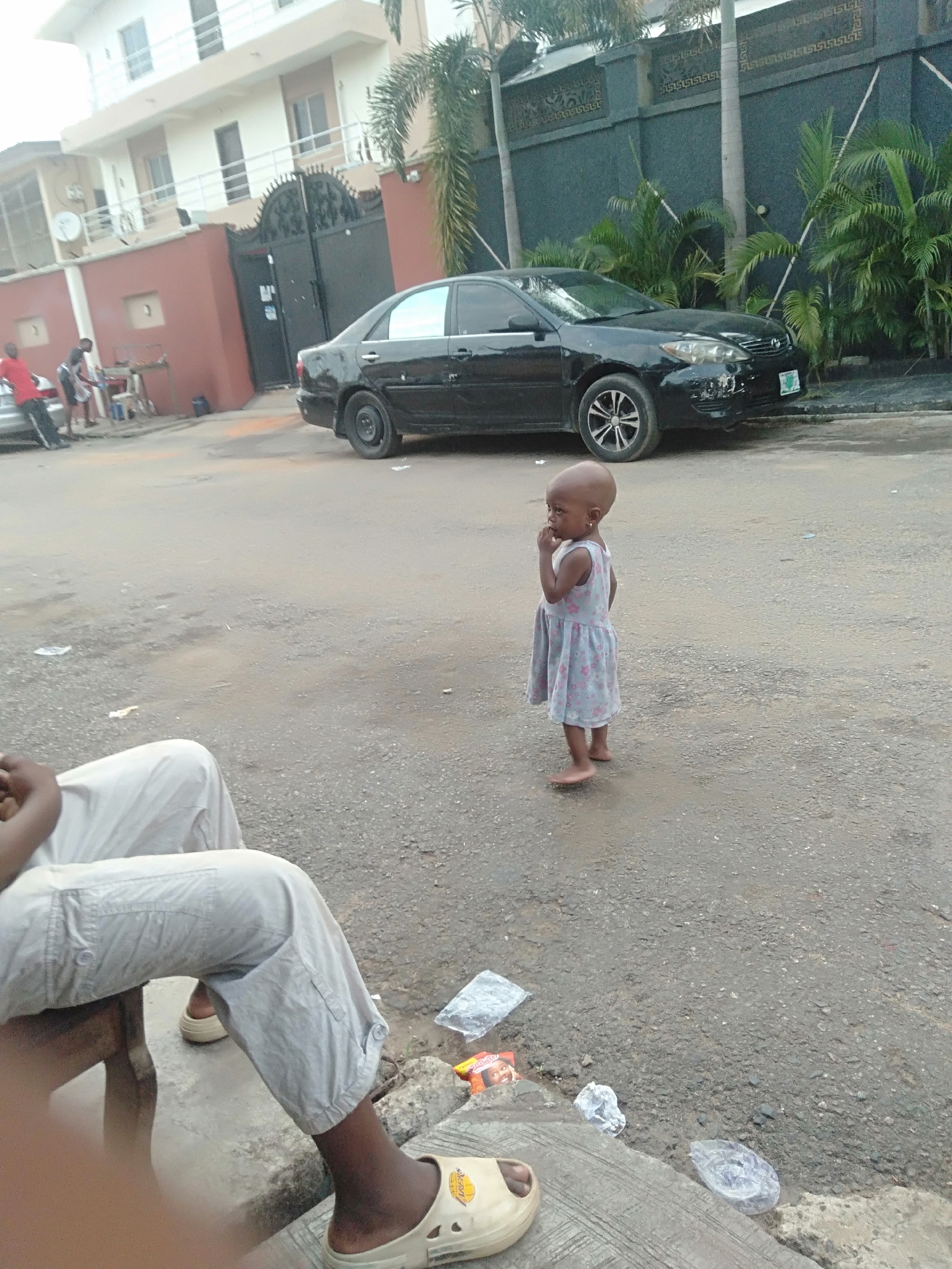 A young child stands barefoot on a paved street in an urban setting. The child wears a light-colored floral dress and gazes to the side with a thoughtful expression. In the background, a black sedan is parked near a building with a dark fence. Palm trees are visible behind the fence. To the left of the child, an individual is seated, wearing light-colored pants and sandals. The street is littered with debris. In the distance, people are standing near another vehicle. The location is in the city of Unknown, Unknown.