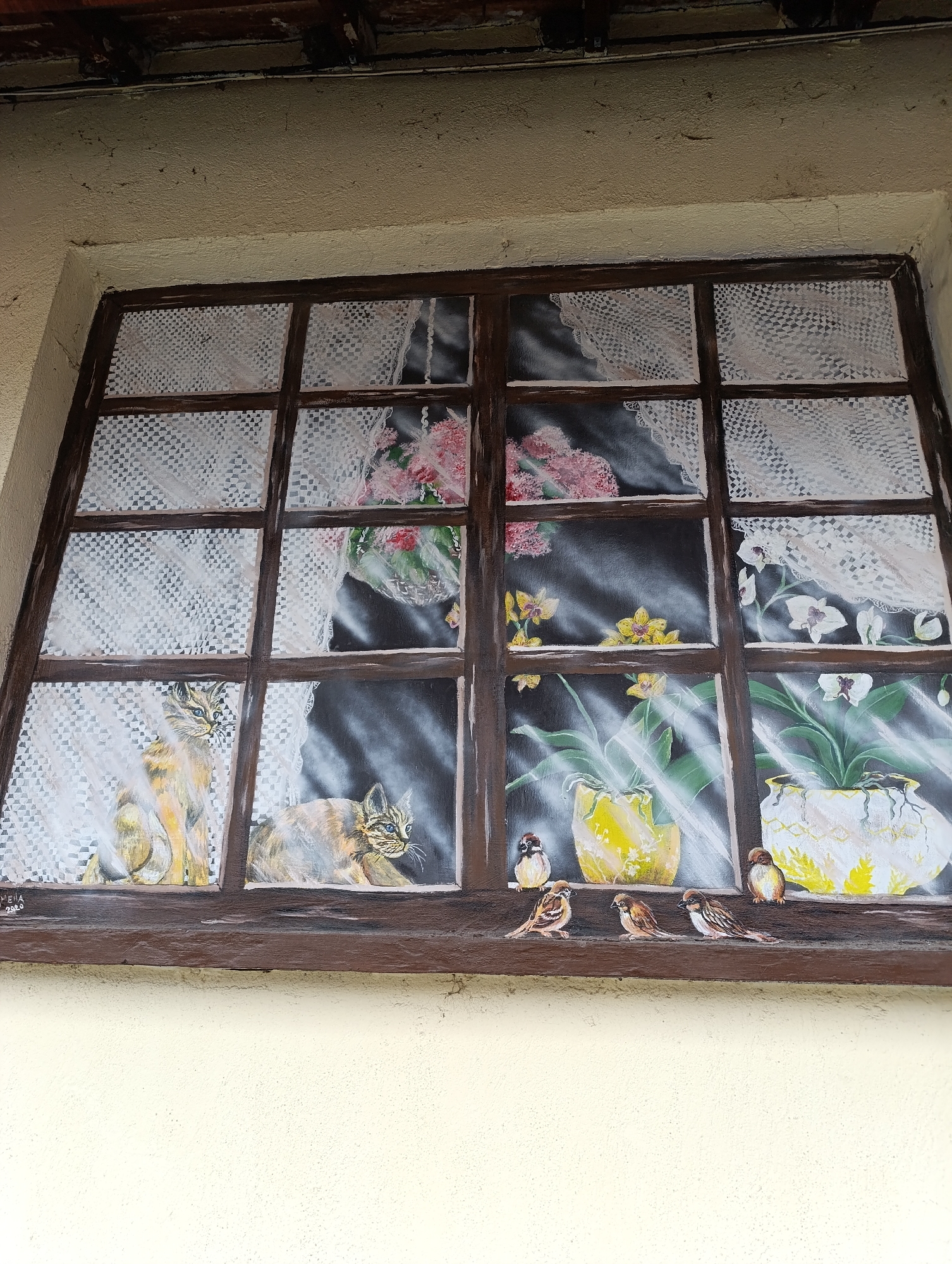 The image depicts a mural painted on a building in Haskovo, Bulgaria. The mural is painted to resemble a multi-paned window with a dark brown frame and a ledge at the bottom. The window panes depict a scene of cats looking out of the window. In the window are potted plants and flowers and lace curtains. On the window ledge are six small sparrows. The building itself appears to be light yellow.