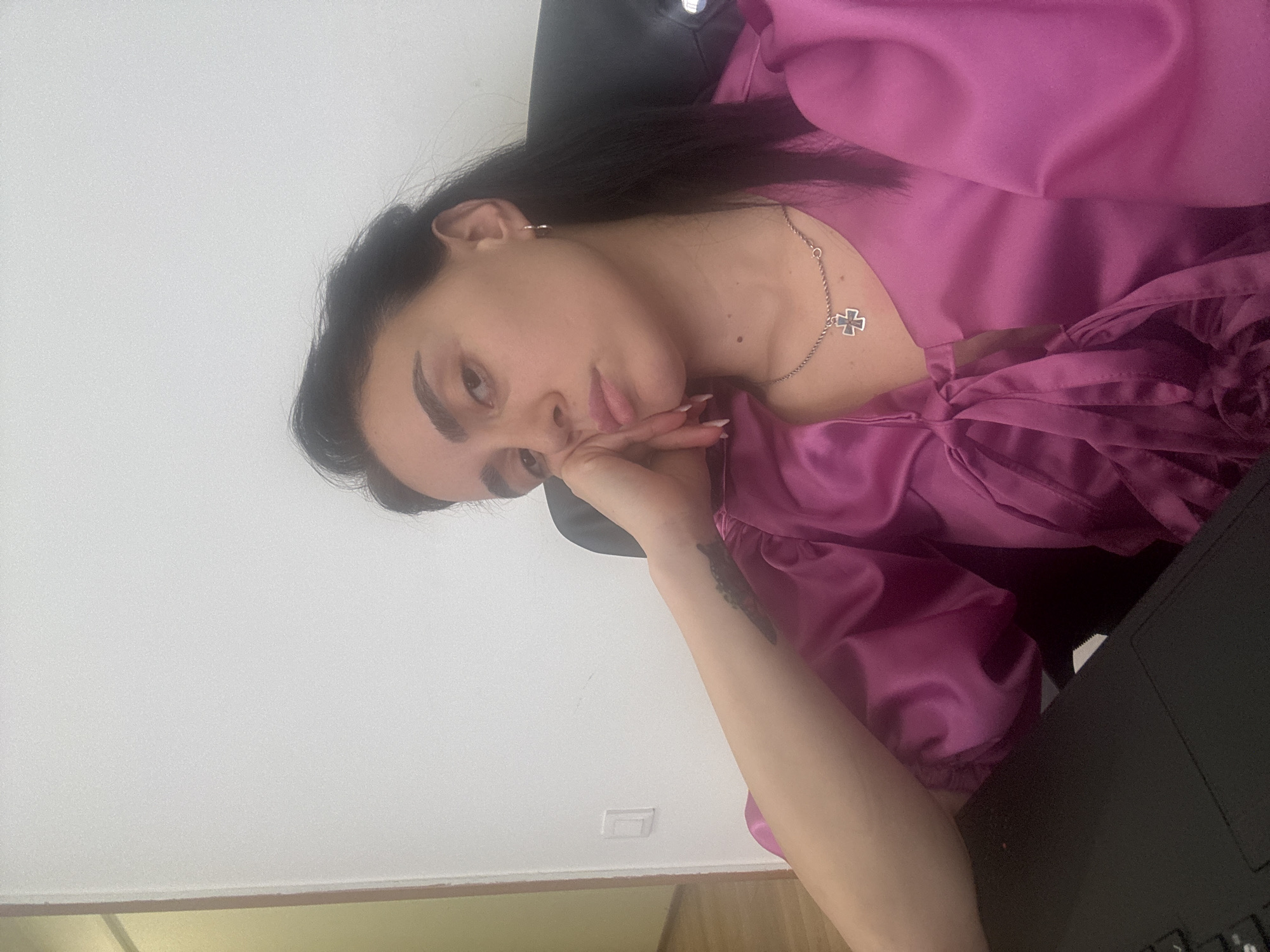 A woman with dark hair, wearing a pink silk top and silver cross necklace, is seated in a black chair in front of a laptop. Her left arm is raised with her hand supporting her head. A tattoo is visible on her left arm. The background consists of a white wall. Based on the image alone, the location in Tbilisi, Georgia cannot be determined.