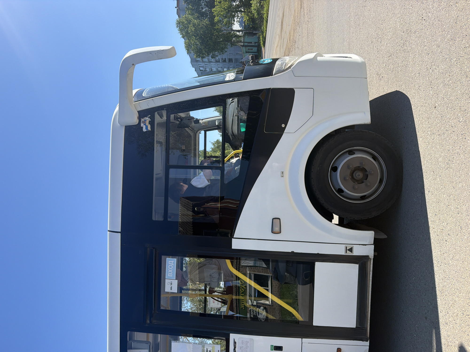 The image shows the side of a white bus parked on an asphalt surface. The bus has dark tinted windows and a white side mirror. A yellow handrail is visible through the windows. On the window, there is a sign with text and icons. The sky is clear and blue, suggesting it is daytime with good weather. In the background, there are trees and a building. The image is taken from a low angle, focusing on the bus.