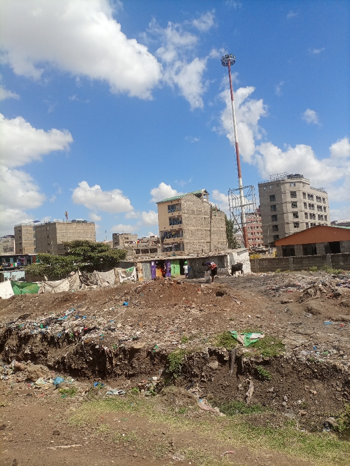 A cityscape is visible under a bright blue sky with scattered clouds. There are several buildings of varying heights and designs, and a tall red and white tower is prominent. In the foreground, there is a ditch filled with garbage and debris. Two people, possibly tending to animals, stand amidst the shacks in front of buildings. The area seems impoverished.