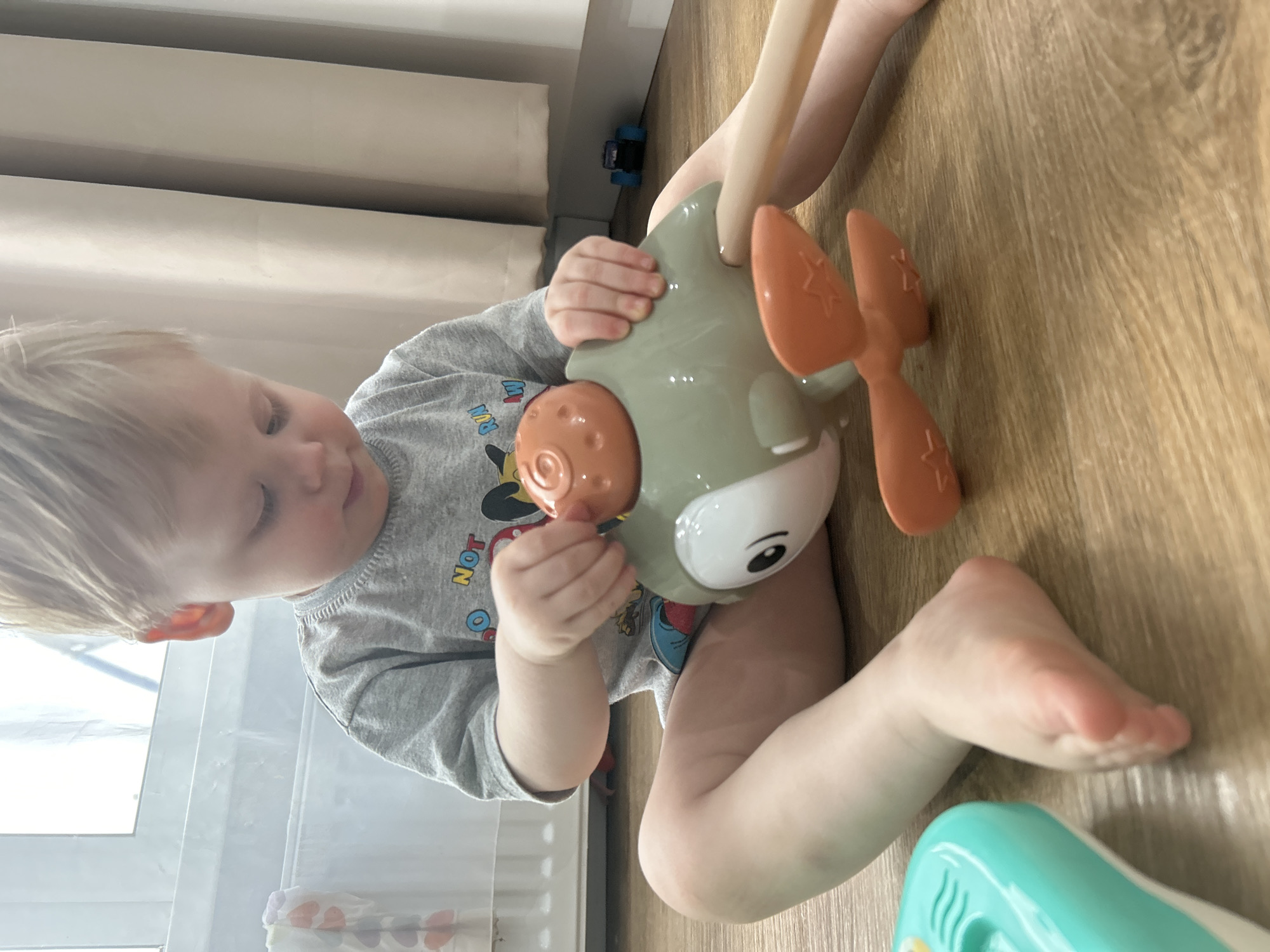 An indoor, eye-level shot, originally rotated clockwise, captures a young child intently playing on a light wooden floor in Chaikovsky, Russia. The child, likely a toddler with short blonde hair, is wearing a light grey t-shirt with a colorful graphic partially visible, including the word "NOT" and what appears to be a cartoon character's ear. They are deeply engrossed in playing with a light green/grey toy airplane, which has prominent orange propellers and a cartoon-like face on its front. The child's hands are wrapped around the toy, suggesting active engagement. Their eyes are cast downwards, indicating concentration.
In the background, a light-colored curtain or blind is visible, along with a small blue toy car tucked under some furniture. Another teal and white toy is partially visible in the bottom right corner of the frame. The setting appears to be a child's play area or living space, illuminated by bright ambient daylight, though the time of day is not precisely discernible. The overall mood is calm and playful, focusing on the child's interaction with their toys.