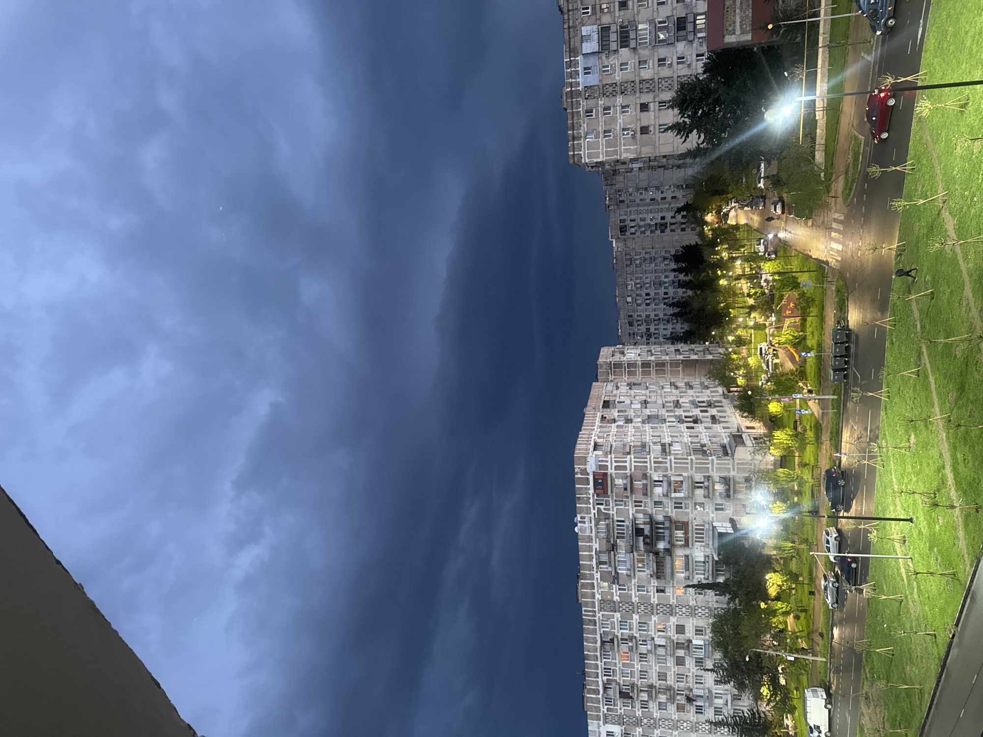 The image presents a view from an elevated position, possibly a balcony or a high-rise building, of a cityscape at twilight or early evening. In the center, dark storm clouds dominate the sky, with a faint crescent moon visible amidst the clouds. On either side, tall residential buildings are seen, their facades showing numerous windows and balconies.
On the right side of the image, a road runs alongside a green area with trees and streetlights. Cars are parked along the road and are also driving. The image depicts an urban setting, and the dense housing suggests a densely populated area. It is Tbilisi, Georgia.