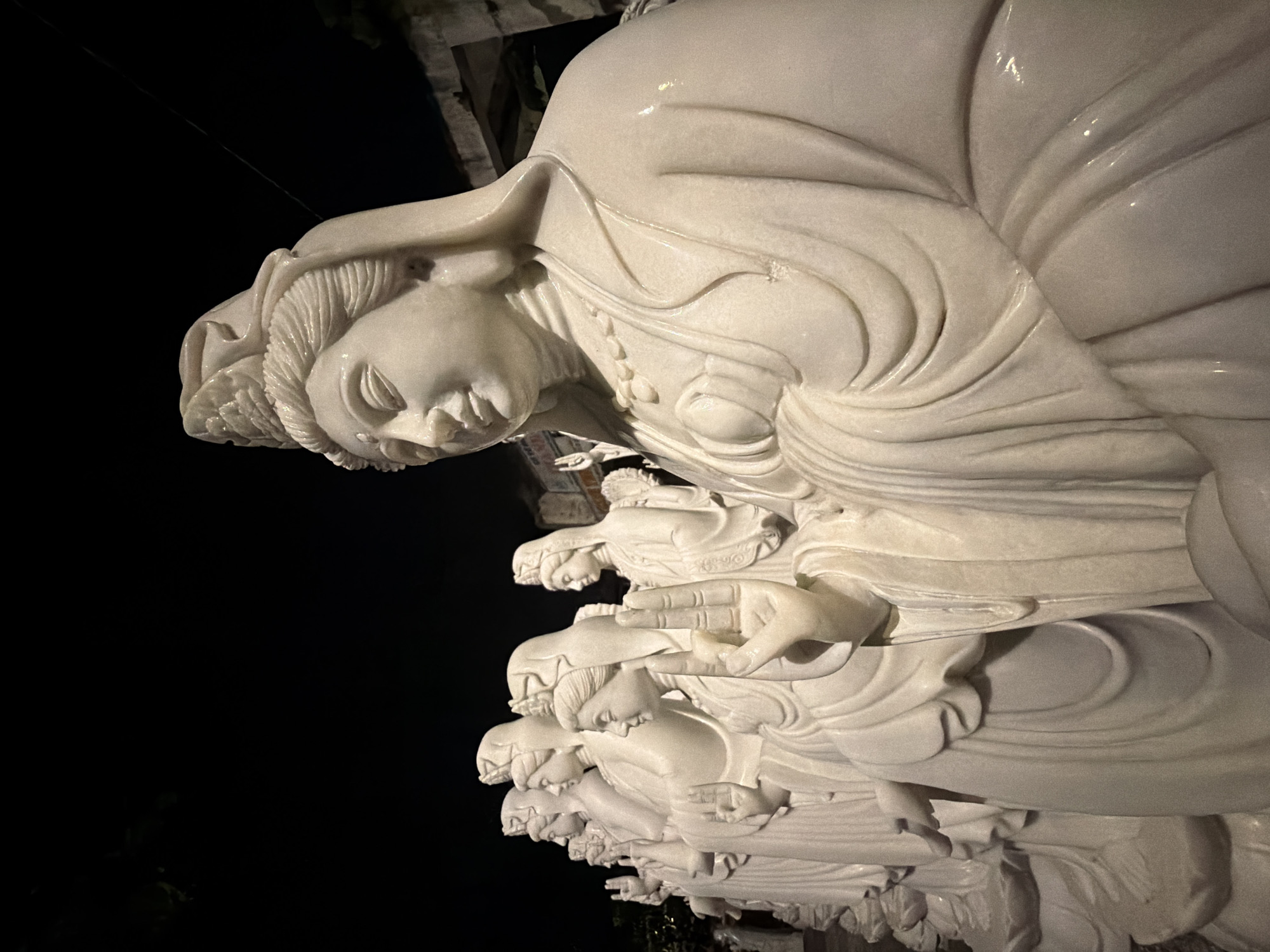 A low-angle, color photograph shows a series of white marble statues depicting women wearing traditional Vietnamese dress and head coverings. The statues are arranged in a linear, stacked formation, with each figure kneeling or reclining in a similar pose. The figures have serene facial expressions and intricately carved details on their clothing and hair. The background is predominantly black, suggesting the photograph was taken at night. The statues are part of the Marble Mountains, a cluster of limestone and marble hills located south of Đà Nẵng, Vietnam. The Marble Mountains are known for their caves, temples, and sculptures, and are a popular tourist destination.