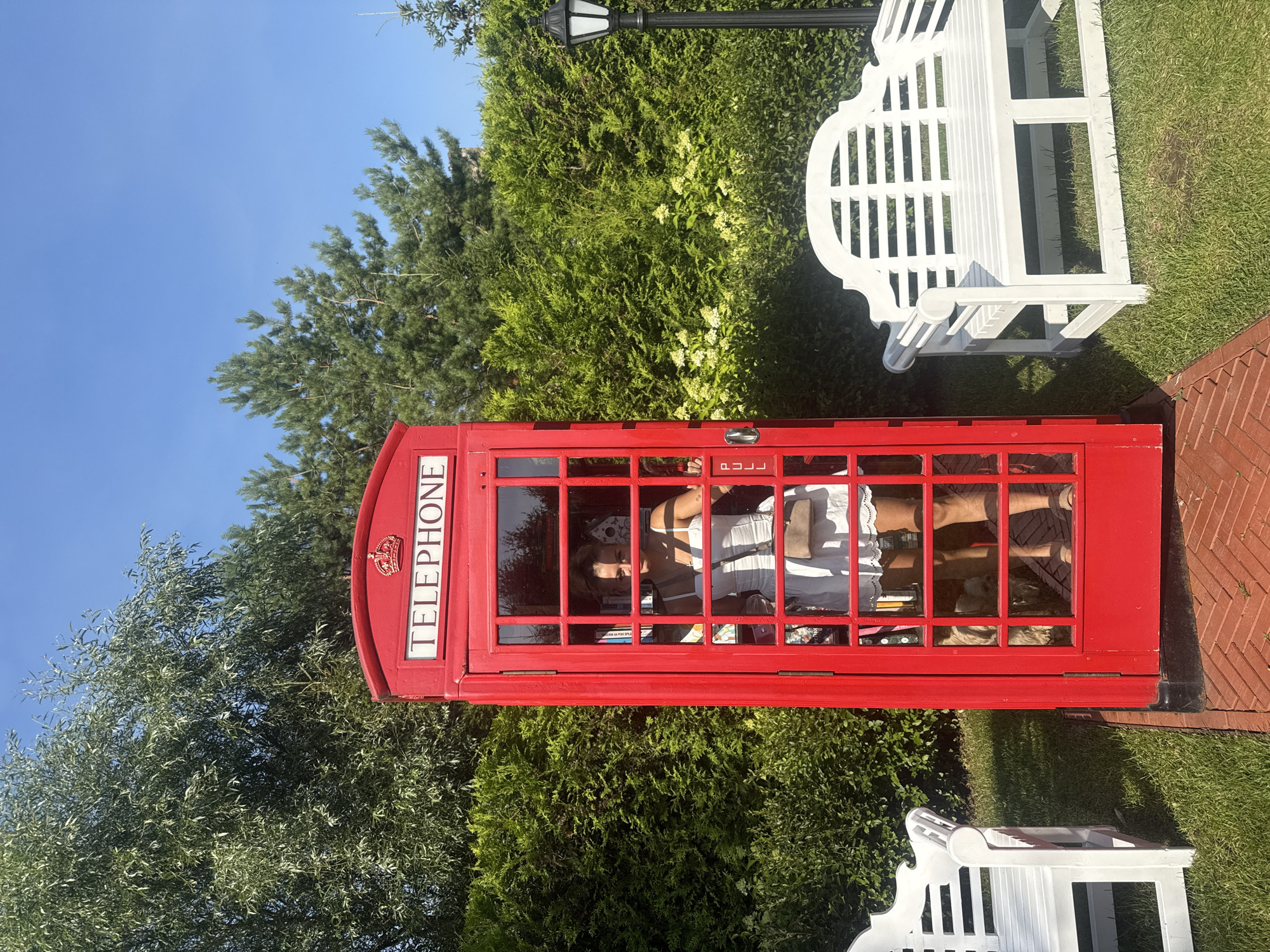 A woman in a white dress stands inside a red telephone booth in a garden setting. The telephone booth is a classic British design with "TELEPHONE" written across the top. The woman is reflected in the glass, and a beige purse is slung over her shoulder. Two white benches flank the telephone booth, one to the left and one to the right. Behind the booth and benches are manicured green hedges, tall trees, and a clear blue sky. A black lamppost stands to the right of the booth. The ground in front of the booth is paved with red bricks, and the rest of the area is covered in green grass. The image is taken in the city of Seltso, Russia, at a garden that appears to be a recreational area or a themed attraction.
