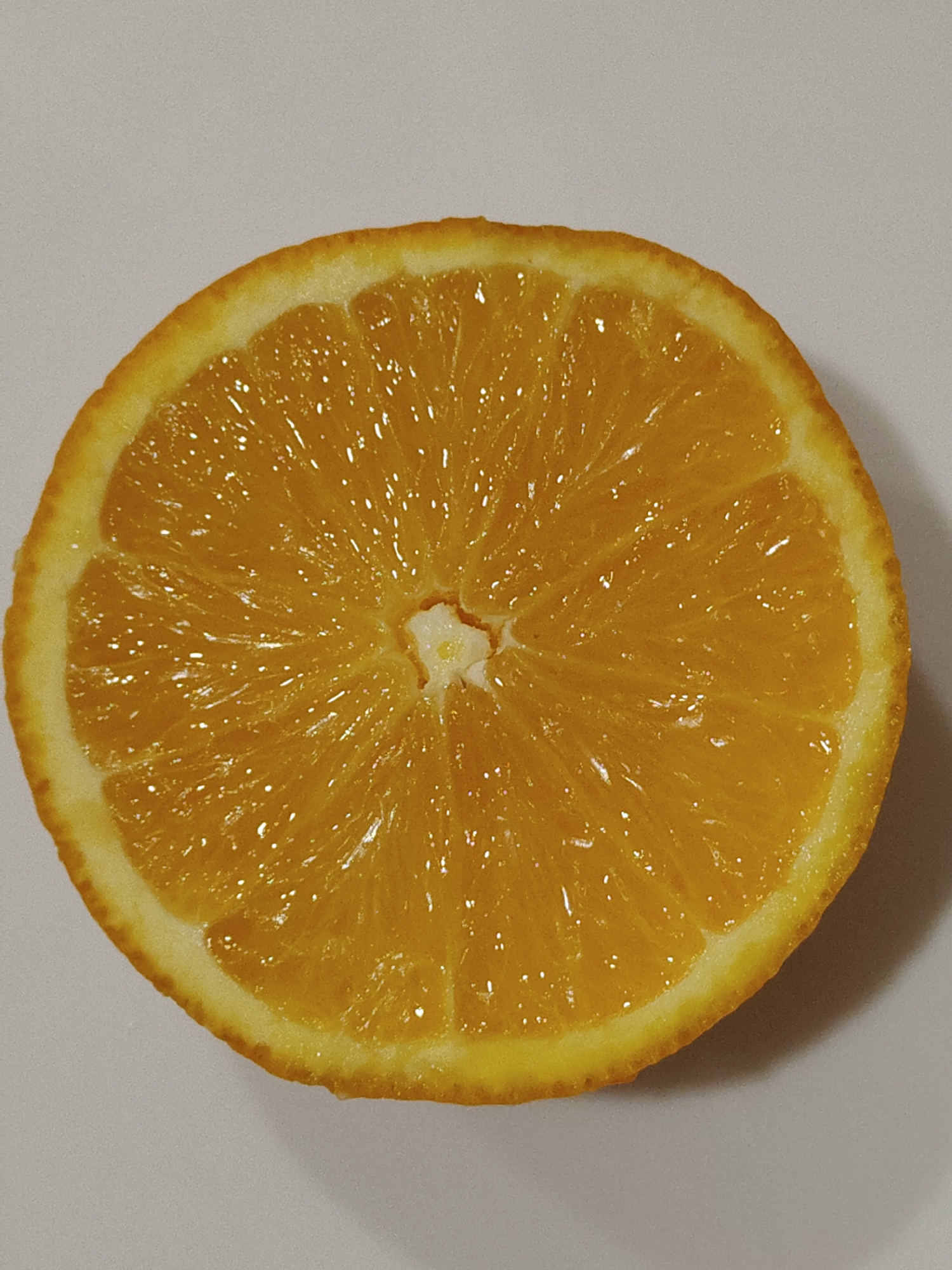 A cross-section of an orange fruit is centered on a neutral, likely white or light grey, surface. The orange displays its vibrant orange segments radiating from the central core, with visible glistening juice sacs. The rind is a darker shade of orange and forms the outer edge of the slice.