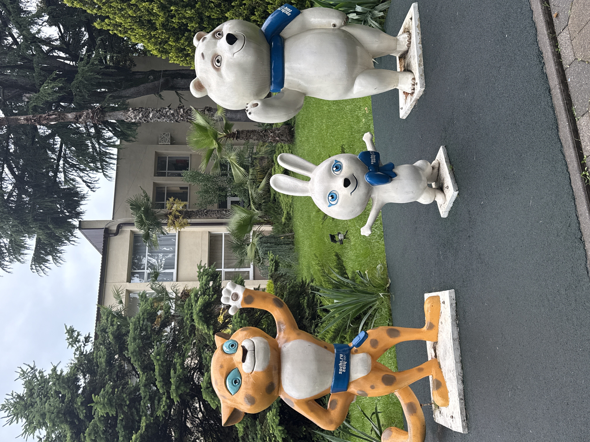 The image depicts three statues of the mascots from the 2014 Winter Olympics in Sochi, Russia. From foreground to background: a tan/brown leopard, a white rabbit, and a white bear, all with blue neck scarves. Each statue is standing on a white platform on a green, black and grey surface. Behind the statues is a building with windows and a beige exterior, surrounded by trees and greenery. The sky is overcast.