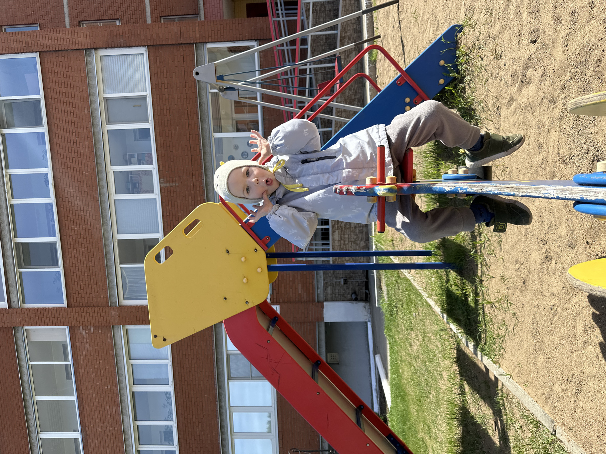 A child is on playground equipment next to a building with brick facing in what appears to be Chaikovsky, Russia. The child is wearing a gray coat, gray pants, green shoes, and a gray hat with small yellow decorations. The playground equipment is primarily red, yellow, and blue. The building is a multi-story structure with several windows. The playground is on a surface of sand and grass. A second piece of playground equipment is visible in the background. The child is looking at the camera, with his mouth open, and holding his hands up.