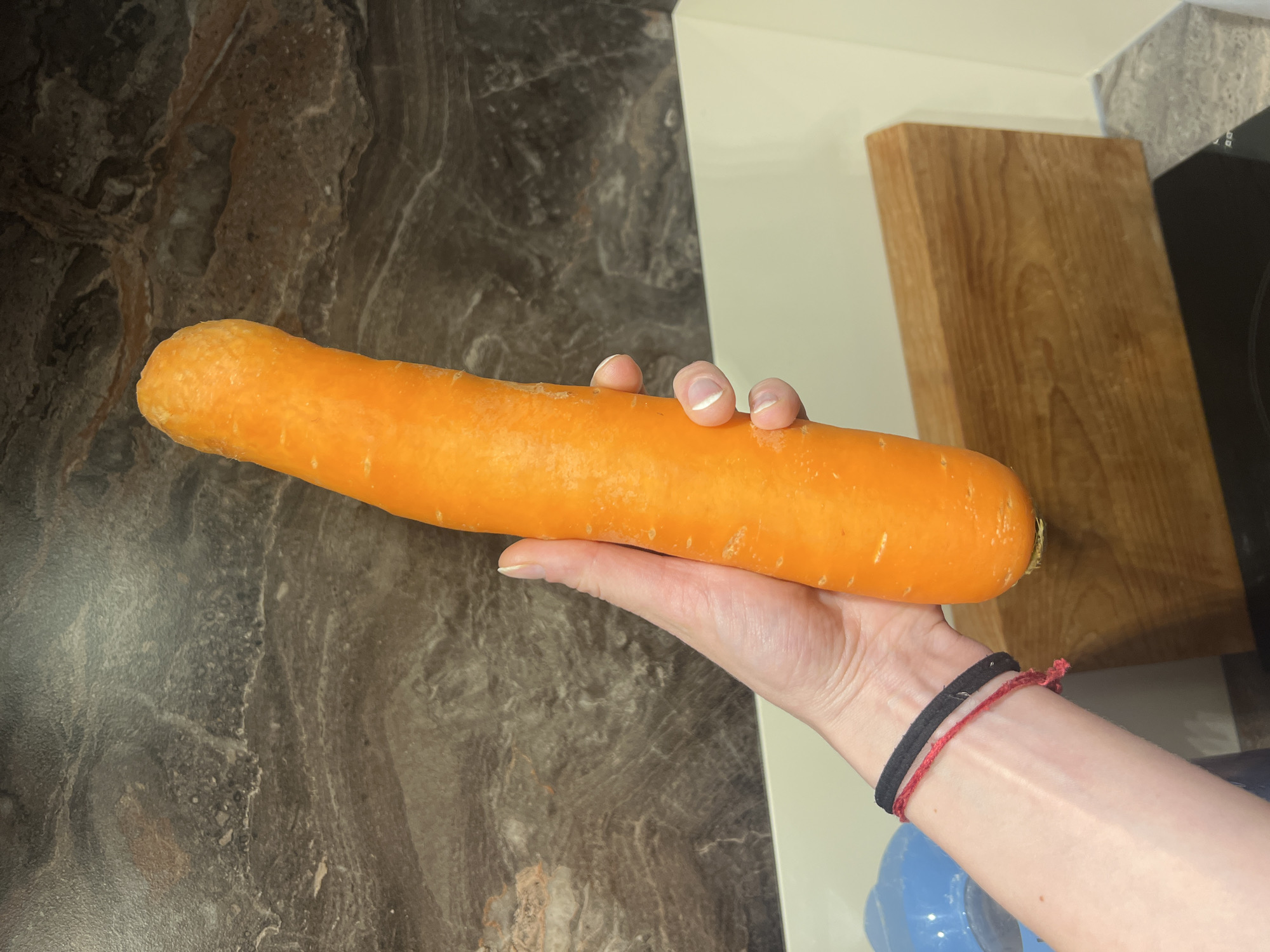 A hand holds a large, peeled carrot against a kitchen countertop. The countertop has a dark, marbled pattern. Behind the hand and carrot, there is a light-colored wooden cutting board near a black appliance. The arm holding the carrot has two wristbands, one black and one red. The setting does not provide enough information to pinpoint the location to Budva, Montenegro.