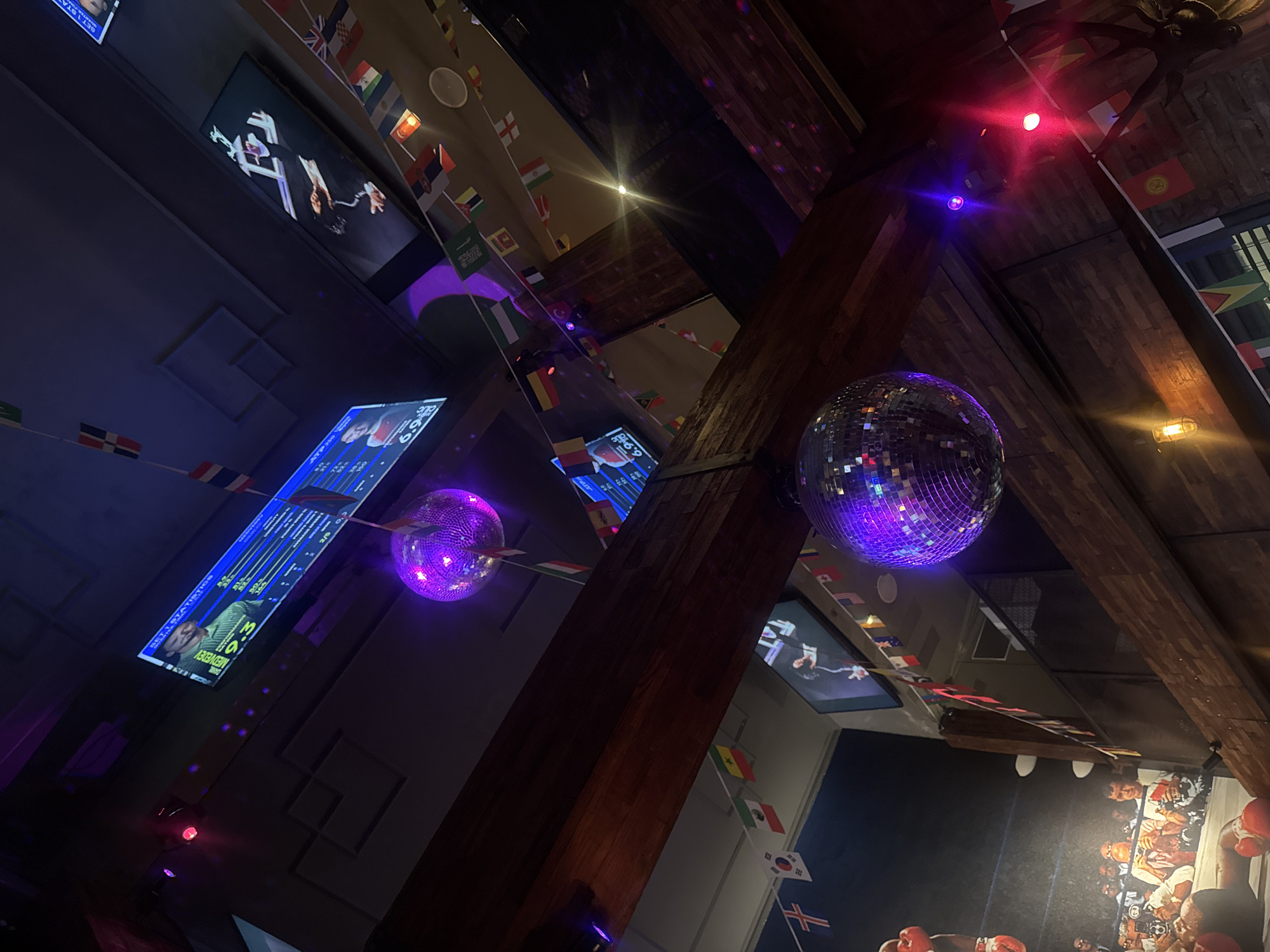 The image depicts the interior of a dimly lit establishment, possibly a sports bar or club, located in North Kuta, Indonesia. The scene is dominated by a wooden beam running diagonally across the frame, supporting the ceiling. Two large, purple disco balls hang from the ceiling, reflecting light and creating a festive atmosphere.
Several screens are visible. One screen on the left displays a dart game with statistics and player names. Another screen in the upper right corner shows a still image of a person in a suit, possibly a performer or commentator. A third screen, partially visible in the upper center, shows an image of a person with the text "SETI STATISTIC" and "DAVE MEDGORY".
A string of small national flags hangs across the ceiling, suggesting an international theme or a gathering of people from various countries. Flags visible include those of Thailand, the United Kingdom, South Korea, Mexico, Italy, and others.
On the left side of the image, a large mural or print features a boxing scene, with a close-up of a boxer's gloved fist in the foreground and other figures in the background.
The environment appears to be a public space, with brickwork visible on the lower right and wooden paneling on the ceiling. Lighting is a mix of colored spotlights (purple and pink) and a warm incandescent bulb hanging from the wooden beam. The overall impression is one of a lively venue, potentially popular for watching sports or enjoying entertainment.